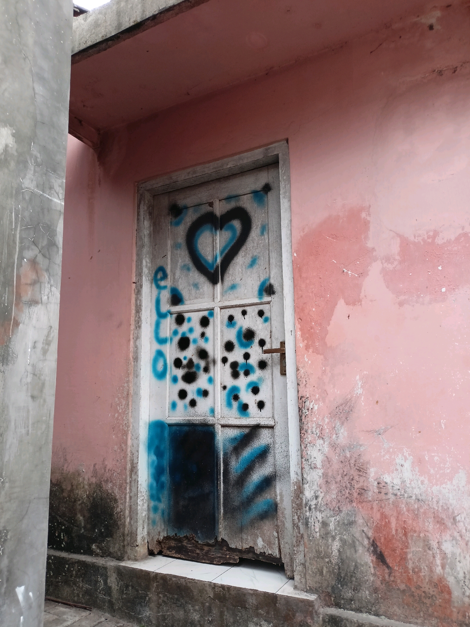 In the city of Payaman, Indonesia, a weathered pink wall is adorned with graffiti. A white door, also covered in spray paint, is set into the wall. The graffiti on the door includes a black heart with a blue outline, blue lettering that appears to say "ECO," and black and blue splatters and stripes. The lower portion of the door is damaged and appears to be rotting. To the left of the door is a concrete pillar. The ground in front of the door is paved with tiles and has some debris on it. The overall impression is one of urban decay and neglect.
