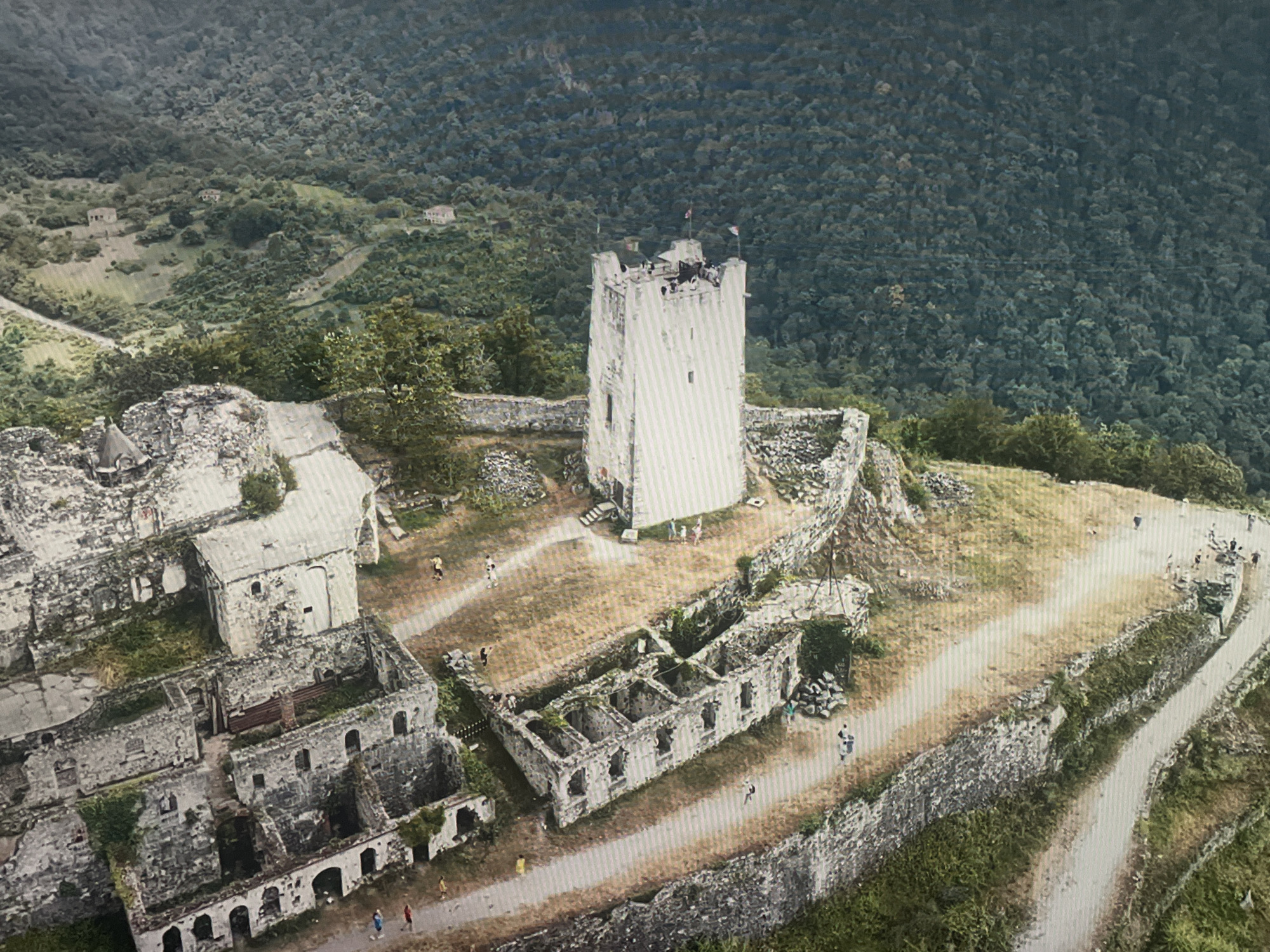 This is an aerial, high-angle shot of the Anacopia Fortress in New Athos, near Sokhumi, Abkhazia. The fortress is situated on a steep, verdant hillside, with dense green forests covering the surrounding mountains.
The central focus is the main structure of the fortress, featuring a prominent, tall, white tower with a crenellated top. The tower has a light-colored facade with visible vertical lines. Surrounding the tower are extensive ruins of stone buildings, many of them partially collapsed, with visible archways and remnants of walls. Some of these structures are overgrown with vegetation. Pathways made of gravel and dirt wind through the complex, connecting different areas.
Several small figures, presumably tourists, are scattered throughout the fortress grounds, walking along the paths and exploring the ruins. Some figures are also visible on top of the main tower. The environment is characterized by rugged terrain, stone structures, and lush natural surroundings. In the background, several houses are visible nestled in the lower-lying areas of the hills. The overall impression is of an ancient historical site set within a dramatic natural landscape.