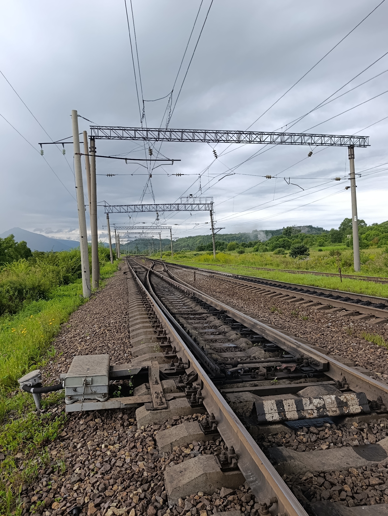 The image shows a set of railroad tracks on a cloudy day in a rural or semi-rural area. The tracks are made of steel rails on concrete sleepers, covered with ballast. Overhead power lines for an electric train are visible, supported by concrete poles. The surrounding environment is green with trees and grass. In the distance, hills and mountains are visible. The scene appears to be the outskirts of the industrial settlement of Londokozavod, Russia, possibly near a railway junction or a switch point. There are no people visible in the image.