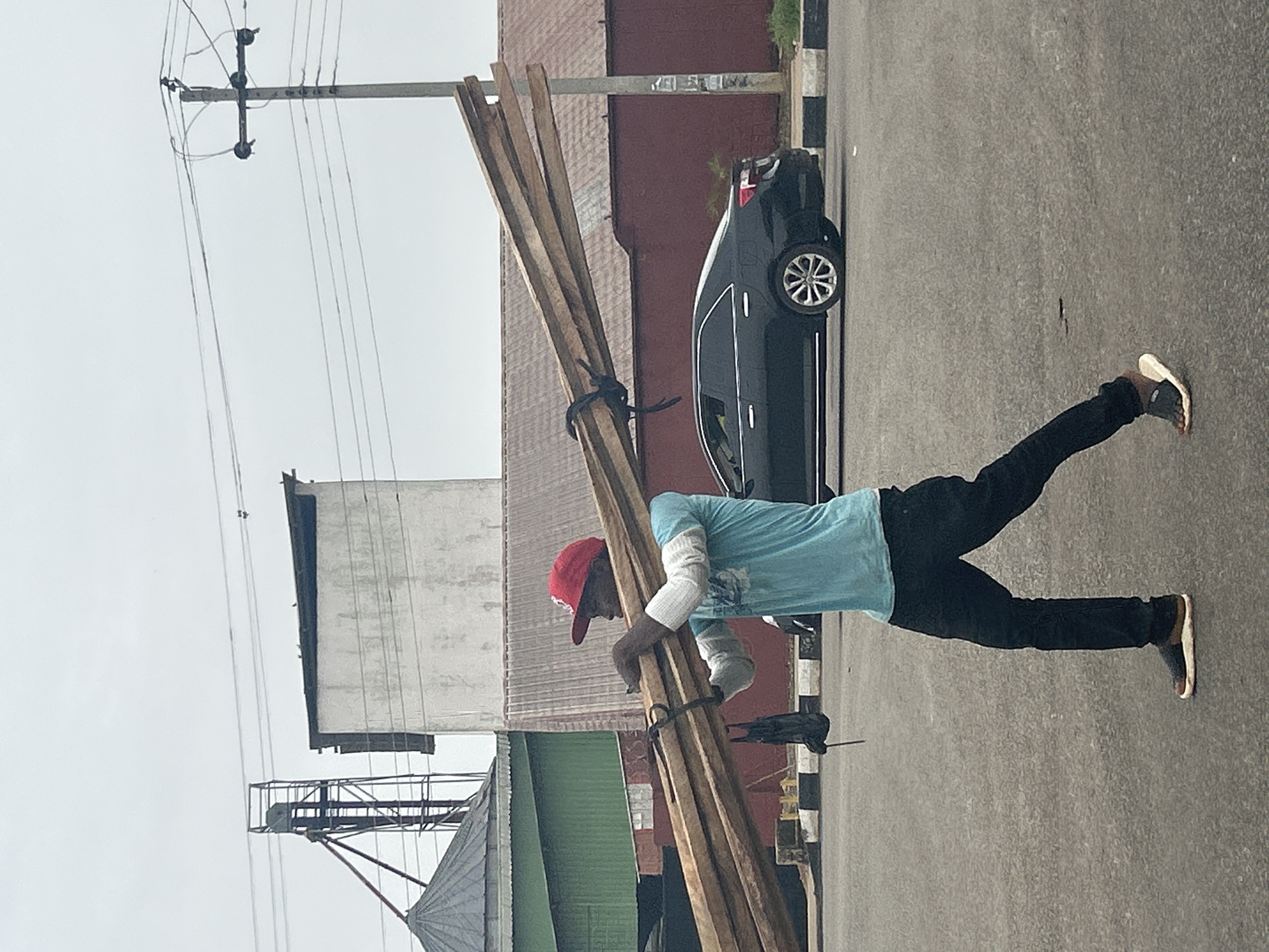 A man in a red cap, light blue t-shirt, and dark pants is carrying a large bundle of wooden planks on his shoulder. He is walking along an asphalt road. Behind him, there is a black car parked on the side of the road, as well as buildings and a utility pole. The photo was taken in Jos, Nigeria.