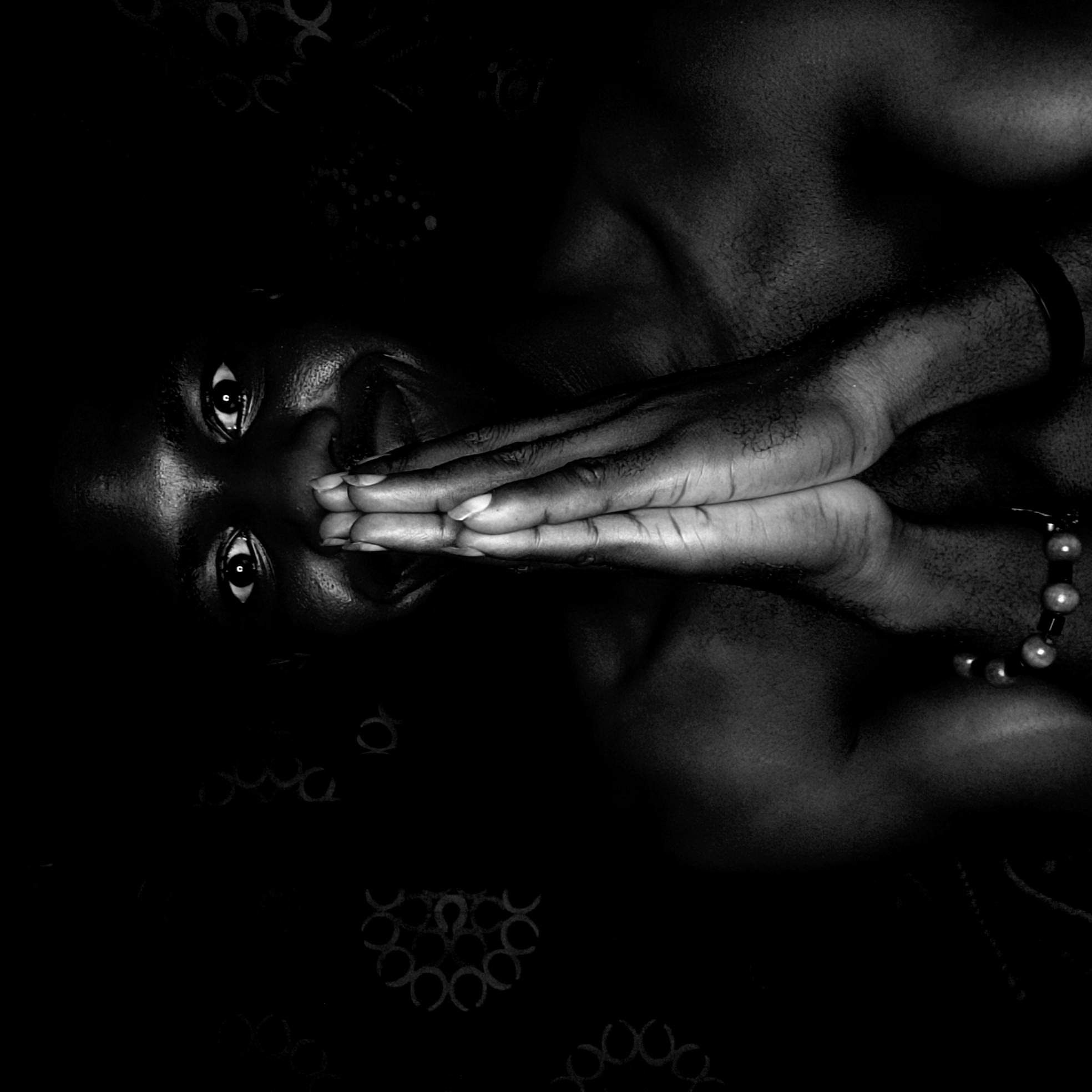 A black and white portrait depicts a person with dark skin, their head tilted to the left. The person's eyes are wide and focused, and their hands are held together, covering their mouth and part of their nose. The lighting is dramatic, with stark contrasts and deep shadows. A bracelet is visible on one wrist. The background is dark and indistinct, with faint patterns. There is no indication that this image depicts the city of Narayi, Nigeria.