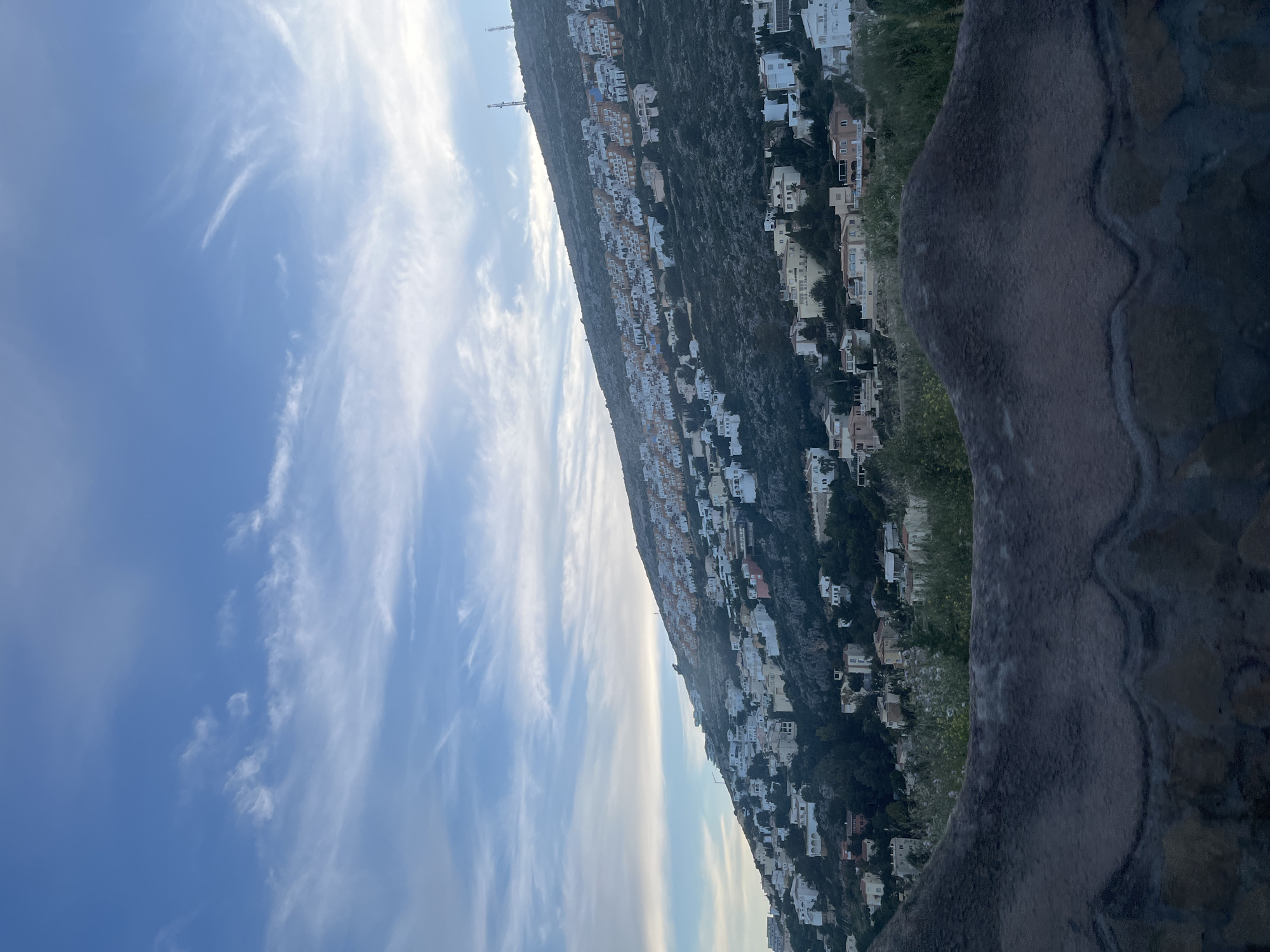 The image shows a hillside in El Poble Nou de Benitatxell / Benitachell, Spain. Many white villas are built on the rocky hillside. The sky is blue with cirrus clouds. The foreground shows a textured, curved structure, possibly a stone wall or part of an architectural element, overlooking the hillside.