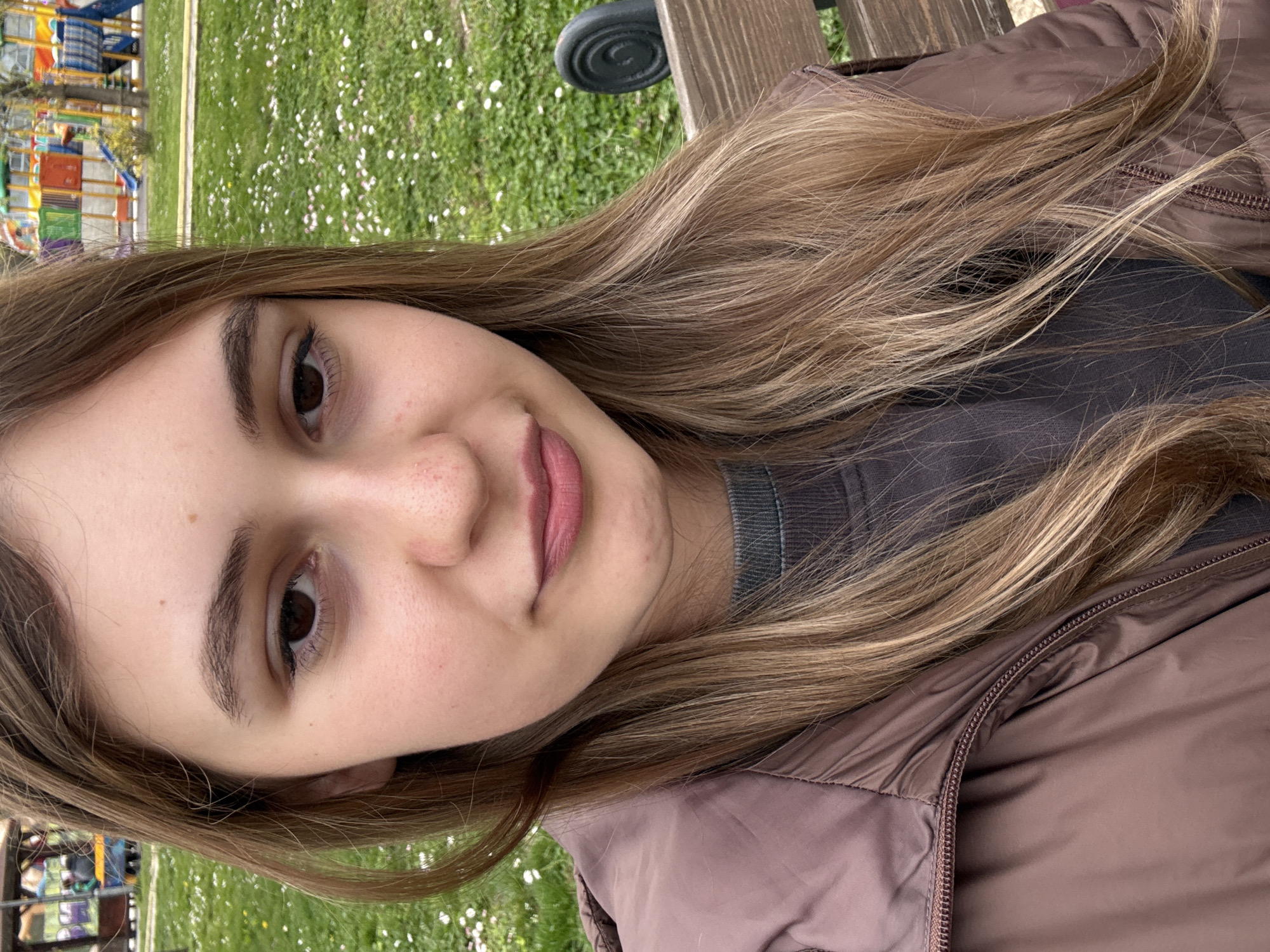 A young woman is featured in a close-up outdoor portrait, set in Panagyurishte, Bulgaria. She has long, wavy hair with shades of brown and lighter highlights, falling over her shoulders and jacket. Her dark brown eyes gaze directly at the camera, and a subtle, closed-mouth smile graces her lips. Visible details on her face include faint freckles or skin texture on her nose and cheeks.
She is wearing a warm, brown or taupe-colored puffer jacket with a visible zipper, and a dark grey or charcoal garment underneath. The setting appears to be a park or public green space during daylight hours. In the background, there's a vibrant green lawn dotted with small white flowers, resembling daisies. To the upper left, a colorful children's playground structure with various slides and climbing sections is partially visible. Behind her right shoulder, parts of a wooden bench and a dark, possibly metallic, outdoor fixture can be seen. The weather appears mild and pleasant. No discernible text is visible in the image.