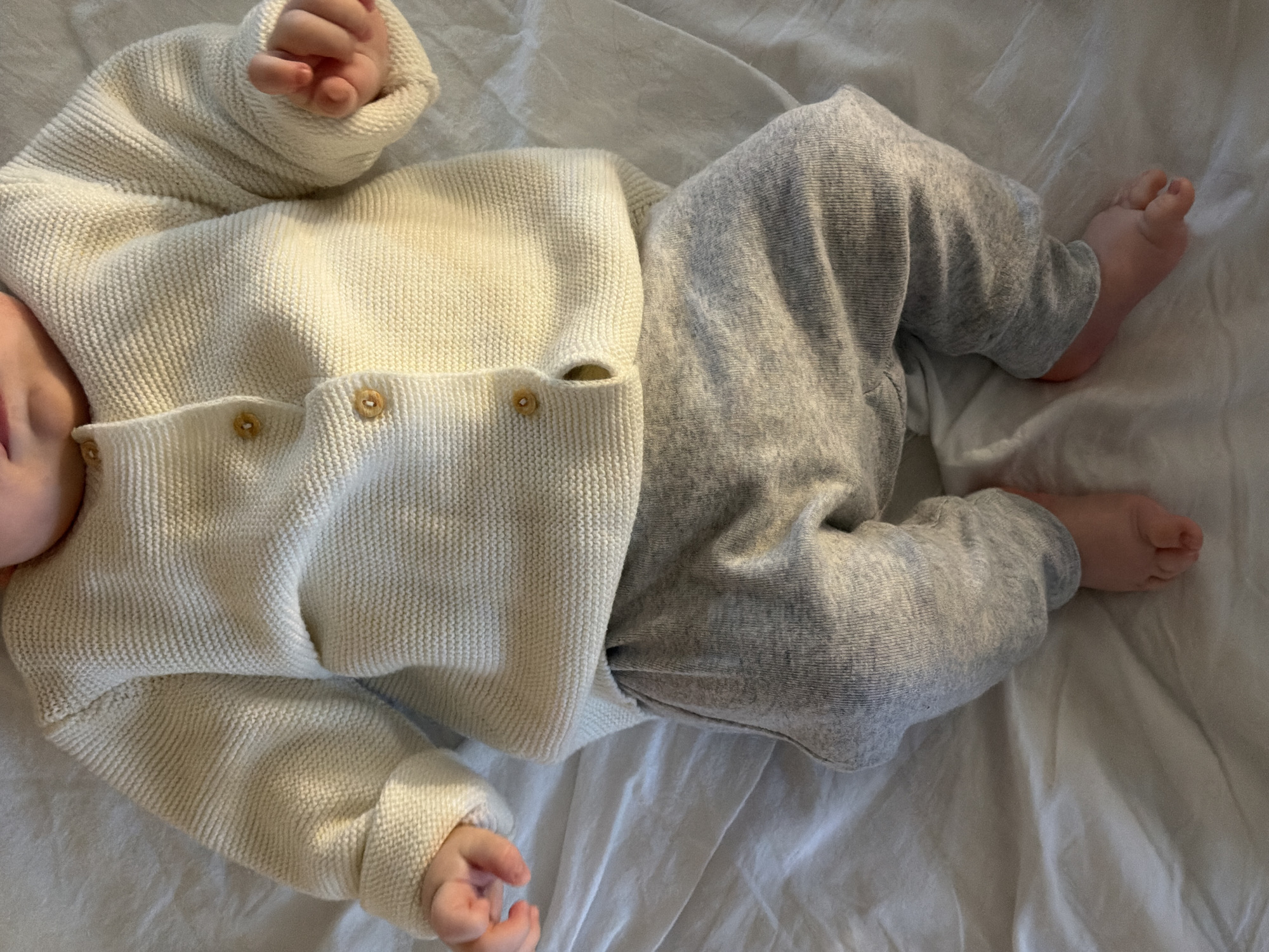 A baby lies on a white, wrinkled surface. The baby is wearing a cream-colored, button-up sweater and grey pants. The baby's hands are visible near the top left of the image, and the baby's bare feet are visible on the right. Only the lower half of the baby's face is visible, near the top left of the image. The surface appears to be a bedsheet. There is no indication that the location is Khimki, Russia.
