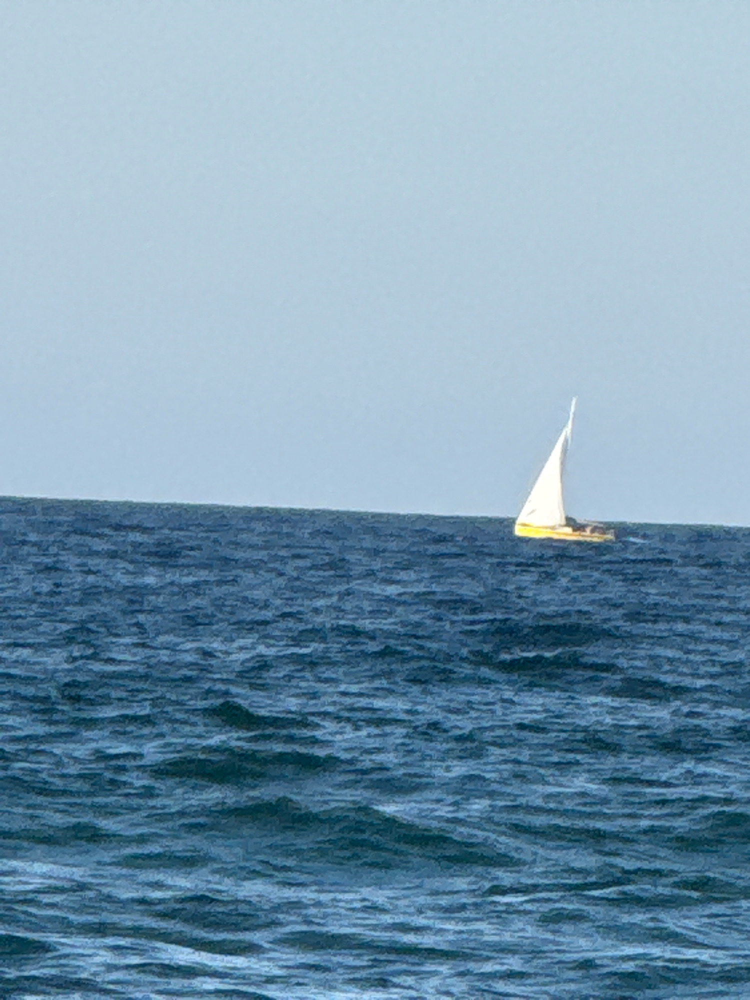 A sailboat with white sails and a yellow hull navigates the deep blue choppy waters of the sea under a clear pale blue sky. The horizon line is visible, dividing the sky and sea. The image appears to be a photograph taken from a distance, possibly from shore or another boat. The location is the sea off the coast of Sozopol, Bulgaria.