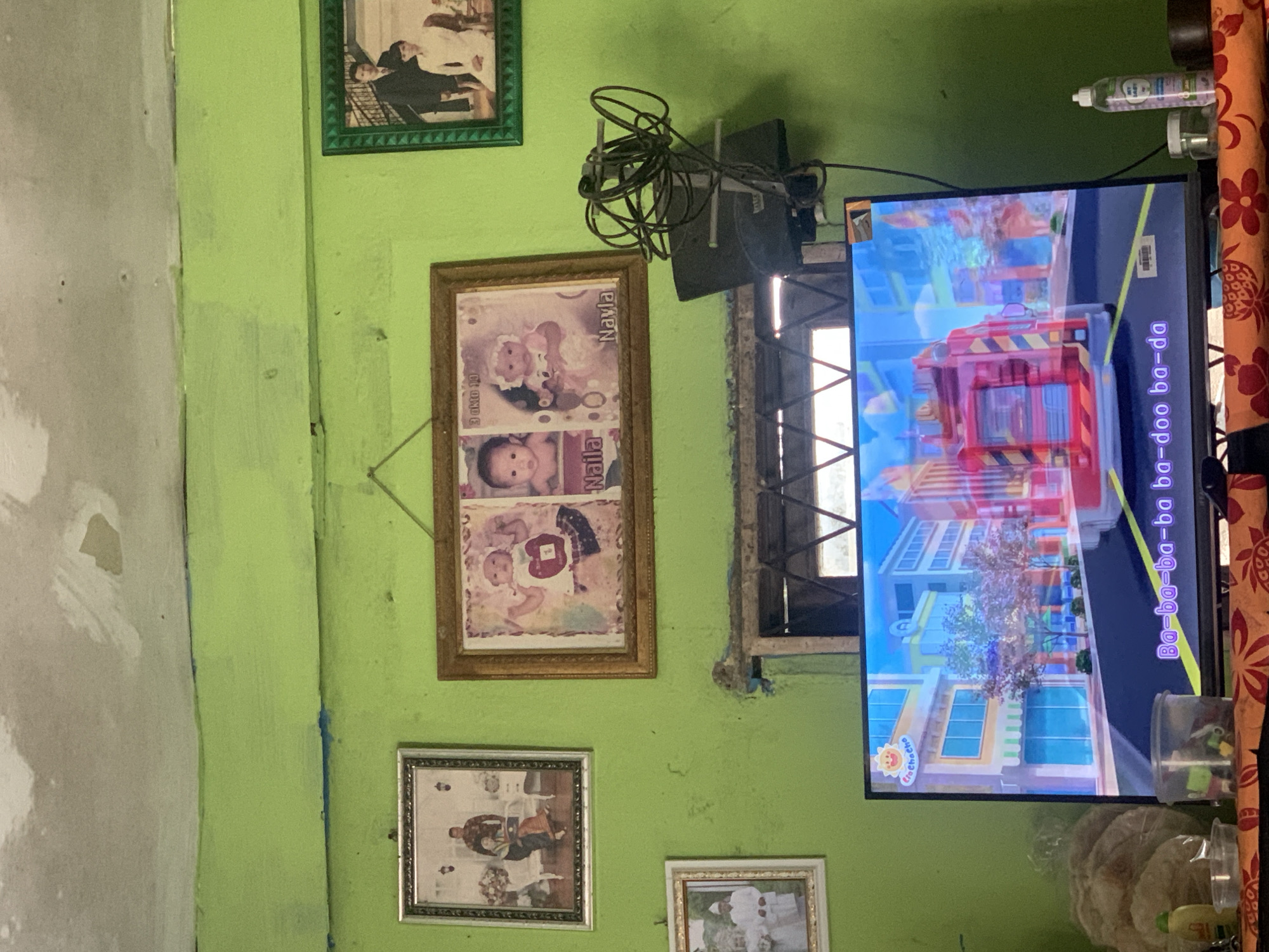 This is a photograph of a television screen displaying a cartoon, in a room with green walls. The television is on a stand, and a wire antenna is mounted above it. On the wall above the television are several framed photographs: a couple dressed formally, two babies with names below them, and a wedding photo. The room appears to be a living space, possibly in Palembang, Indonesia, given the context provided. The overall impression is one of a modestly furnished domestic interior.