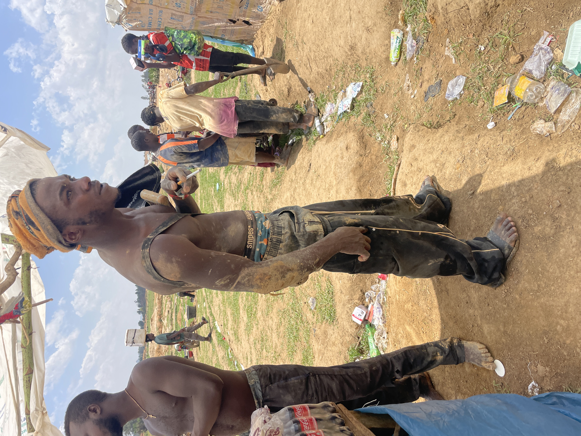 The image depicts several shirtless African men working in a dirty, possibly impoverished, area in what appears to be Maiyanga, Nigeria. The location is littered with trash. One man stands in the foreground, shirtless with dirt on his body and wearing a headwrap. He holds what appears to be a rolled cigarette and is looking away, as other men work behind him. One man is bent over in the lower portion of the image. Other men are visible further in the background, working with shovels and carrying items on their heads. Cement packaging is visible in the upper portion of the image. The environment seems to be an outdoor work site.