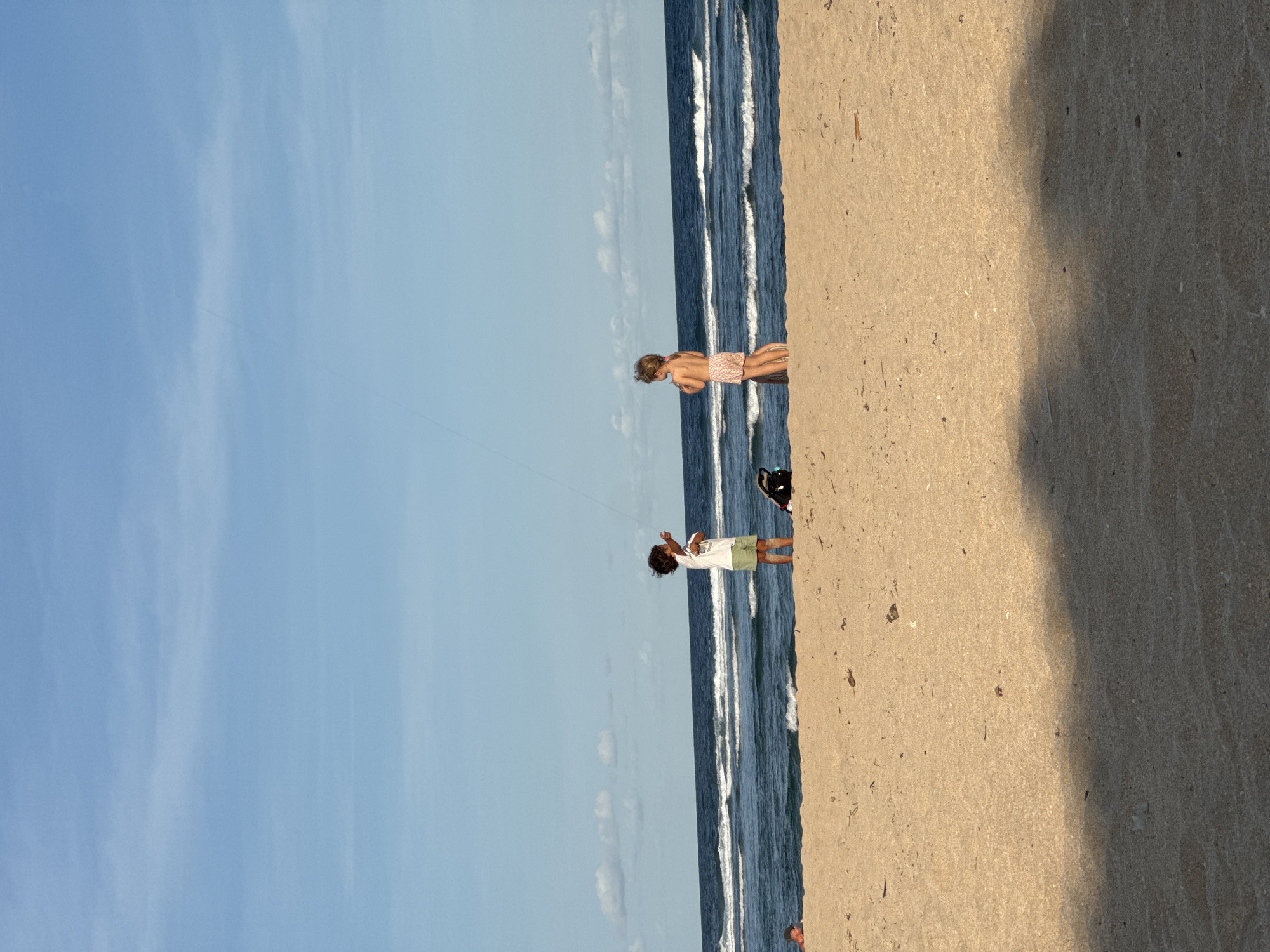 The image depicts a beach scene, possibly in Kuta Selatan, Indonesia. There are two children on the beach. One child, wearing a white shirt and green shorts, is flying a kite. Another child is wearing pink swim trunks, is standing near the shore. A black bag is located on the sand near the water's edge. The sky is blue with scattered clouds. Waves are visible crashing onto the shore. The sand appears light brown.