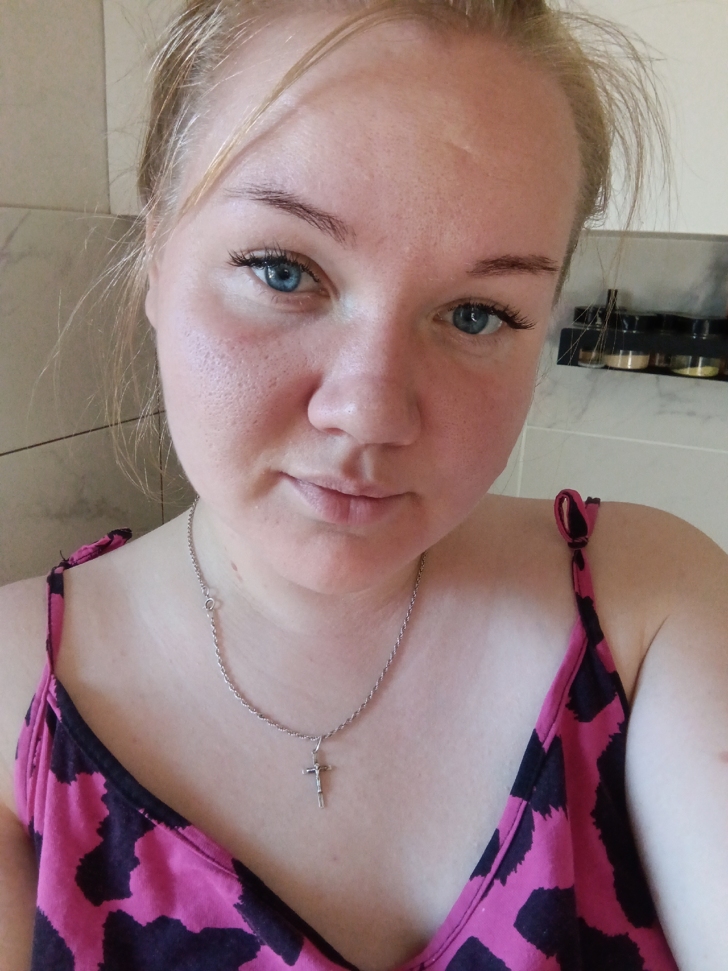 A close-up self-portrait photograph shows a fair-skinned woman with light blonde hair pulled back, wearing a pink and black patterned tank top and a silver necklace with a cross pendant. She has blue eyes and appears to be in a bathroom with a tiled wall visible behind her. There is a shelf with several containers on it in the background. There is no indication this photograph was taken in the city of Фоки, Russia.