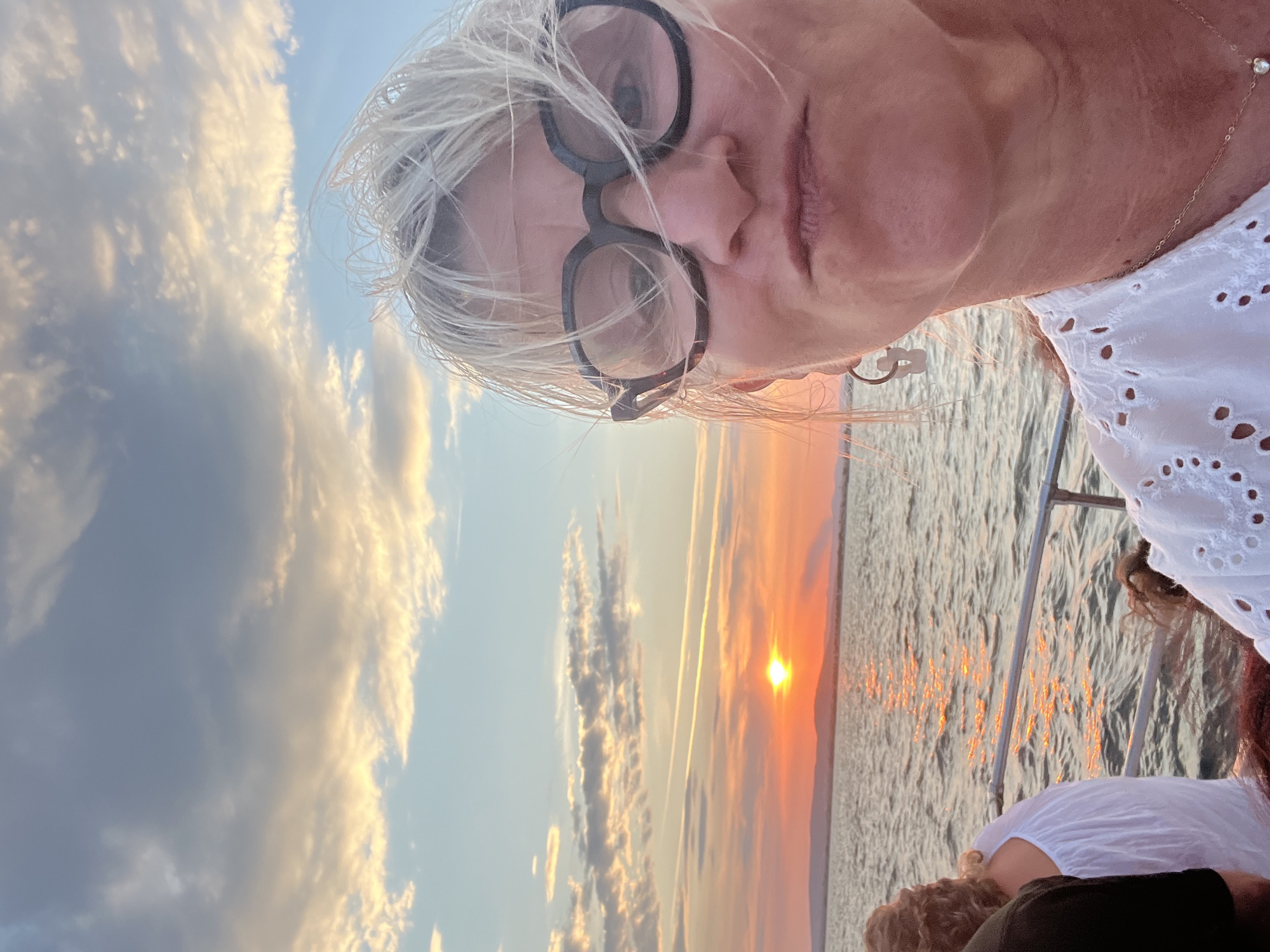 This image captures a selfie taken by a woman with blonde, windswept hair and thick-rimmed glasses. She is on a boat, looking directly at the camera with pursed lips. Behind her, the sun is setting over a body of water, casting a warm glow on the clouds and the rippling surface. The horizon is marked by a distant landmass. Other people are visible on the boat, including a woman in a white top with her back to the camera and another person to the left wearing a dark shirt. The scene appears to be taking place in Palavas-les-Flots, France, given the characteristic coastline and the warm, Mediterranean sunset. The overall atmosphere is relaxed and scenic, suggesting a leisure activity like a sunset cruise.
