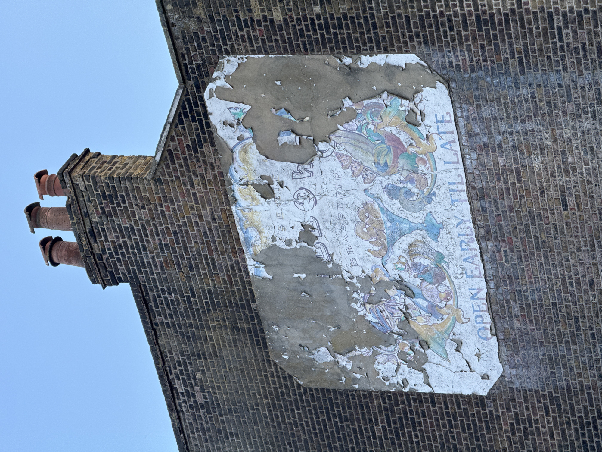 The image shows a brick wall with an old, damaged sign attached to it. The sign, which is mostly peeling away, features some faded illustrations and the words "OPEN EARLY TIL LATE". The wall appears to be part of a building, possibly in London, United Kingdom, given the architectural style.