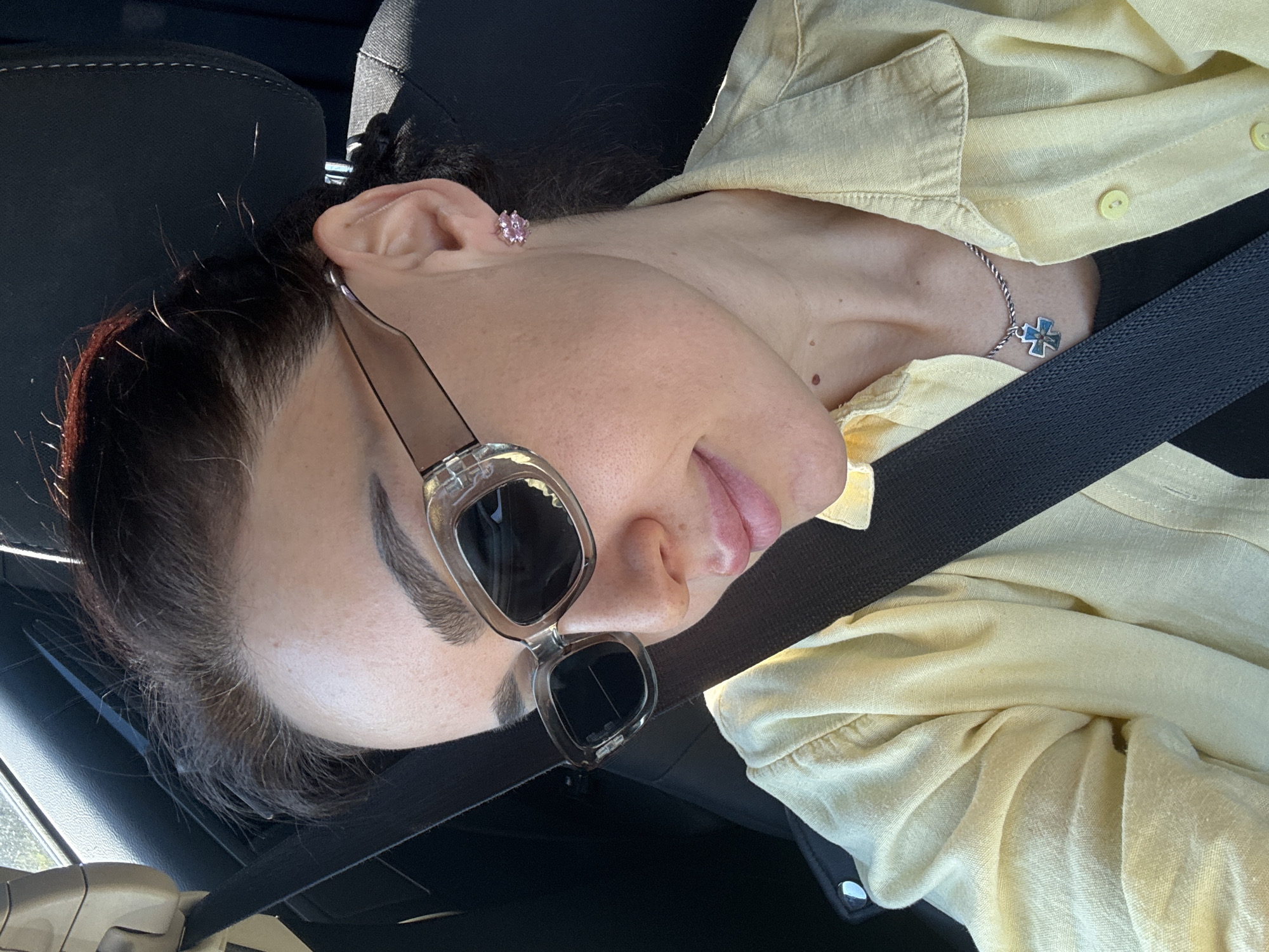 A person wearing sunglasses and a yellow button-down shirt is in a car, secured by a seatbelt. The person is wearing a pink earring and a necklace with a blue cross. The background includes a black car seat and the side window. The location is not discernible from the image.