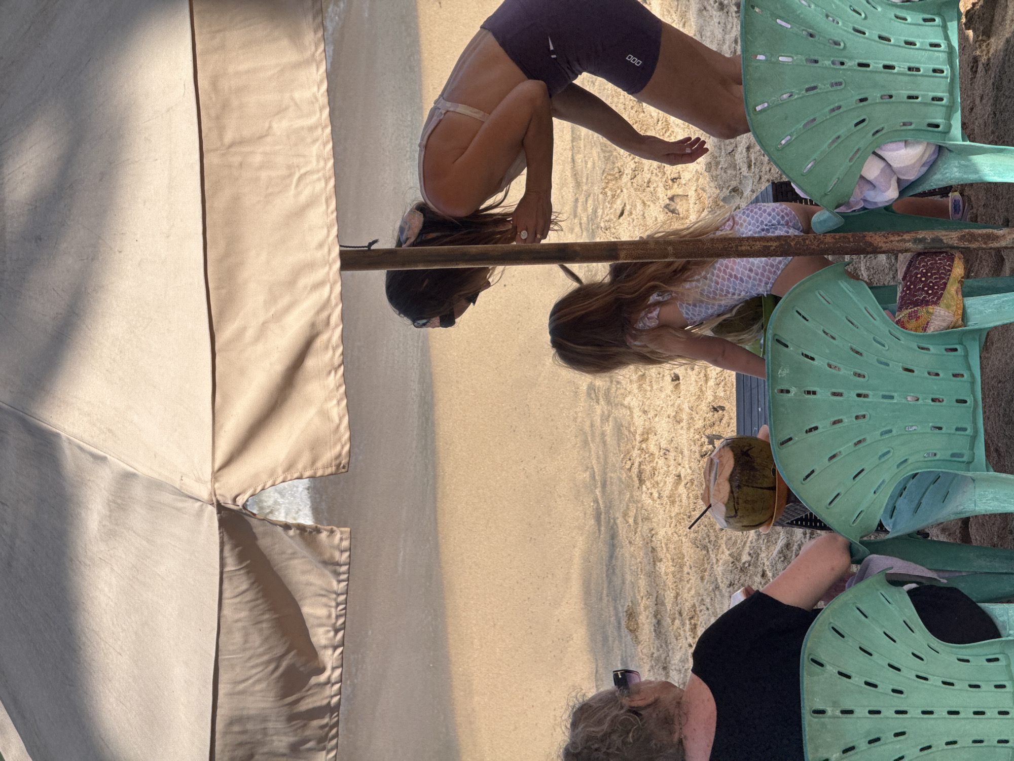 In Kuta, Indonesia, a scene unfolds on a sandy beach, partially shaded by a large, beige umbrella. In the foreground, two teal plastic chairs sit empty, one with a rolled-up towel and what appears to be a patterned sarong tucked into it. A woman with curly, light-colored hair and sunglasses is seated in the left chair, facing away from the camera and towards the water. She is wearing a black top and dark pants.
To the right, a woman in a light-colored bikini top and dark athletic shorts bends over, attending to a young girl. The girl, wearing a colorful swimsuit with a mermaid scale pattern, is also leaning forward, seemingly engaged in an activity at a low table positioned between them and the seated woman. On the table, a whole coconut with a straw is visible. The background shows the golden sand of the beach, with gentle waves of the ocean visible in the distance. The overall lighting suggests a warm, sunny day, with dappled shadows cast by the umbrella.