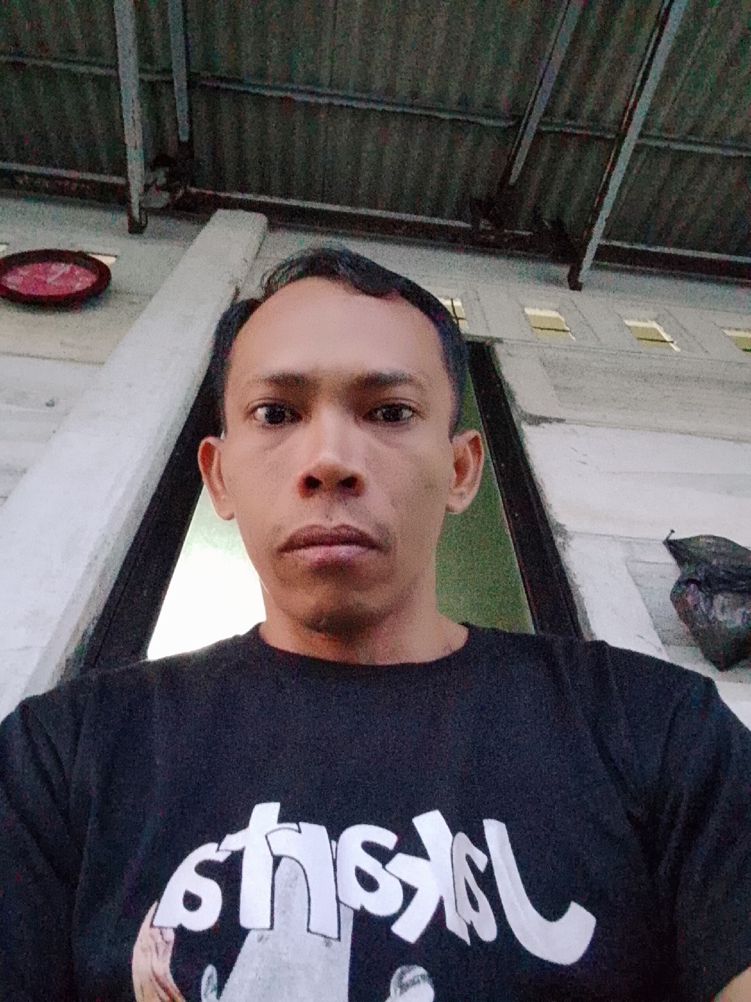 A medium-shot photograph shows a man with short black hair, brown eyes, and a mustache. He is centered in the frame, wearing a black t-shirt. The writing on the t-shirt is white and appears to be upside-down, but it is partially obscured. He has a neutral expression.
The background suggests an outdoor location. The immediate backdrop behind the man is a white structure with a window visible to his right. Above the structure, there is a corrugated metal roof supported by metal bars. In the top left corner of the frame, a small red oval object is mounted on the structure. To the right of the structure, there is what appears to be a black garbage bag. The location is Bukit Ketok, Indonesia.