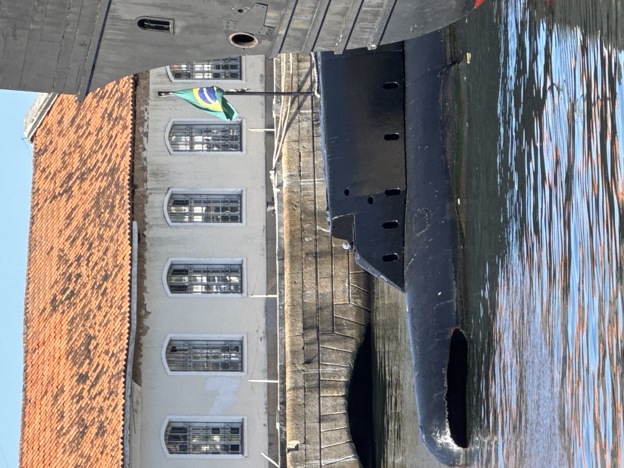 A black submarine is docked in a body of water. In the background, a building with an orange tile roof and arched windows with bars is visible. A Brazilian flag flies from a pole in front of the building. The image was taken in the city of Rio de Janeiro, Brazil.