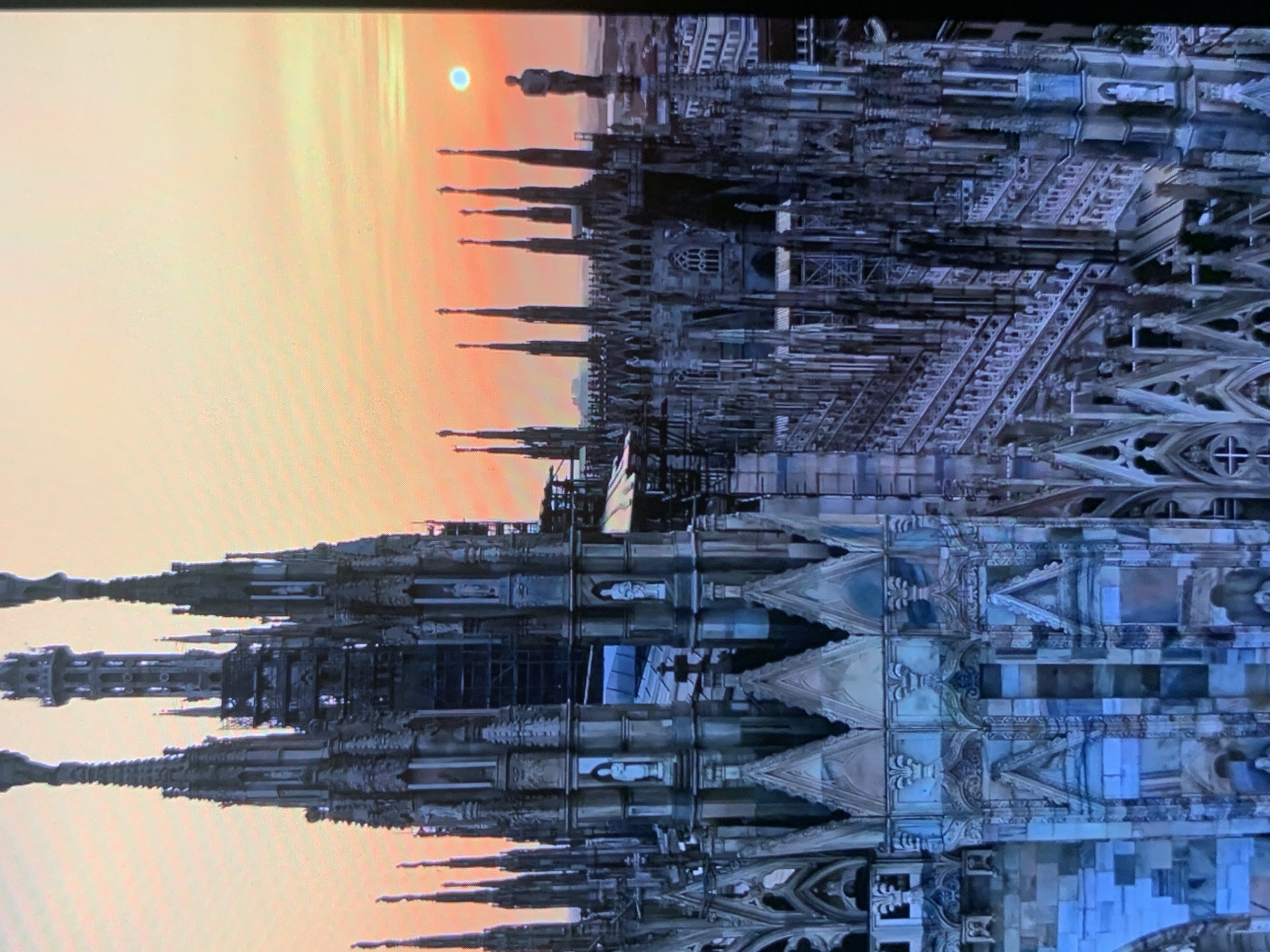 The image shows a panoramic view of a gothic cathedral with intricate spires and architectural details, bathed in the warm glow of a sunset. The sky transitions from a pale yellow near the horizon to a soft orange, with a faint sun visible as a small blue circle. The cathedral's structure is characterized by numerous tall, pointed spires, some adorned with statues. The stonework of the cathedral displays a blend of light and shadow, highlighting its detailed ornamentation and complex design. In the distance, city buildings can be seen extending towards the horizon. The image is a photograph taken from a high vantage point, offering a sweeping perspective of the cathedral and its surroundings during twilight. The location is identified as the city of Stavropol, Russia.