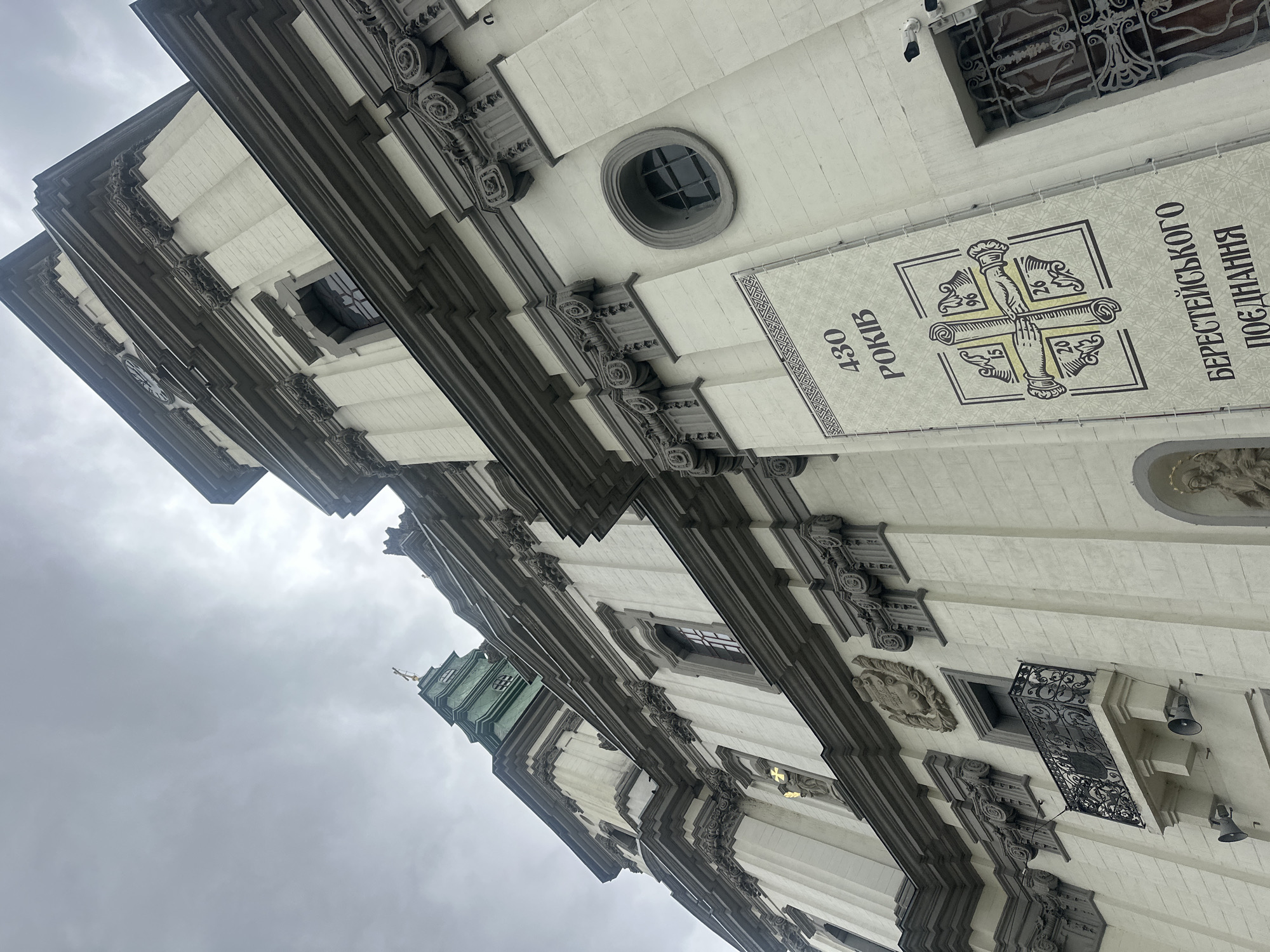 The image shows a low-angle view of the exterior of a large, ornate building, likely a church or cathedral, under a cloudy sky. The architecture features a light-colored facade with dark grey, decorative elements, including columns, moldings, and window frames. A green spire with a cross is visible at the top of the building on the left side.
On the right side of the building, a large banner is displayed. The banner is light-colored with dark text and graphics. The text in Ukrainian appears to read "430 РОКІВ" (430 years) and below that "БЕРЕСТЕЙСЬКОГО ПОЄДНАННЯ" (Union of Brest). The graphic depicts four hands holding scrolls within a square border, with numbers "15", "96", "26", and "20" in the corners, likely representing dates. A security camera is mounted on the wall above and to the right of the banner. A barred window is visible to the right of the banner.
The building also features a balcony with a wrought-iron railing, a small window above it, and two speakers mounted below the balcony. A relief sculpture of a religious figure is visible in an arched niche on the lower right side of the building. The overall impression is of a historic and significant religious or institutional building.
This building is the Ternopil Greek Catholic Cathedral of the Immaculate Conception of the Blessed Virgin Mary, located in Ternopil, Ukraine. The banner commemorates the 430th anniversary of the Union of Brest.