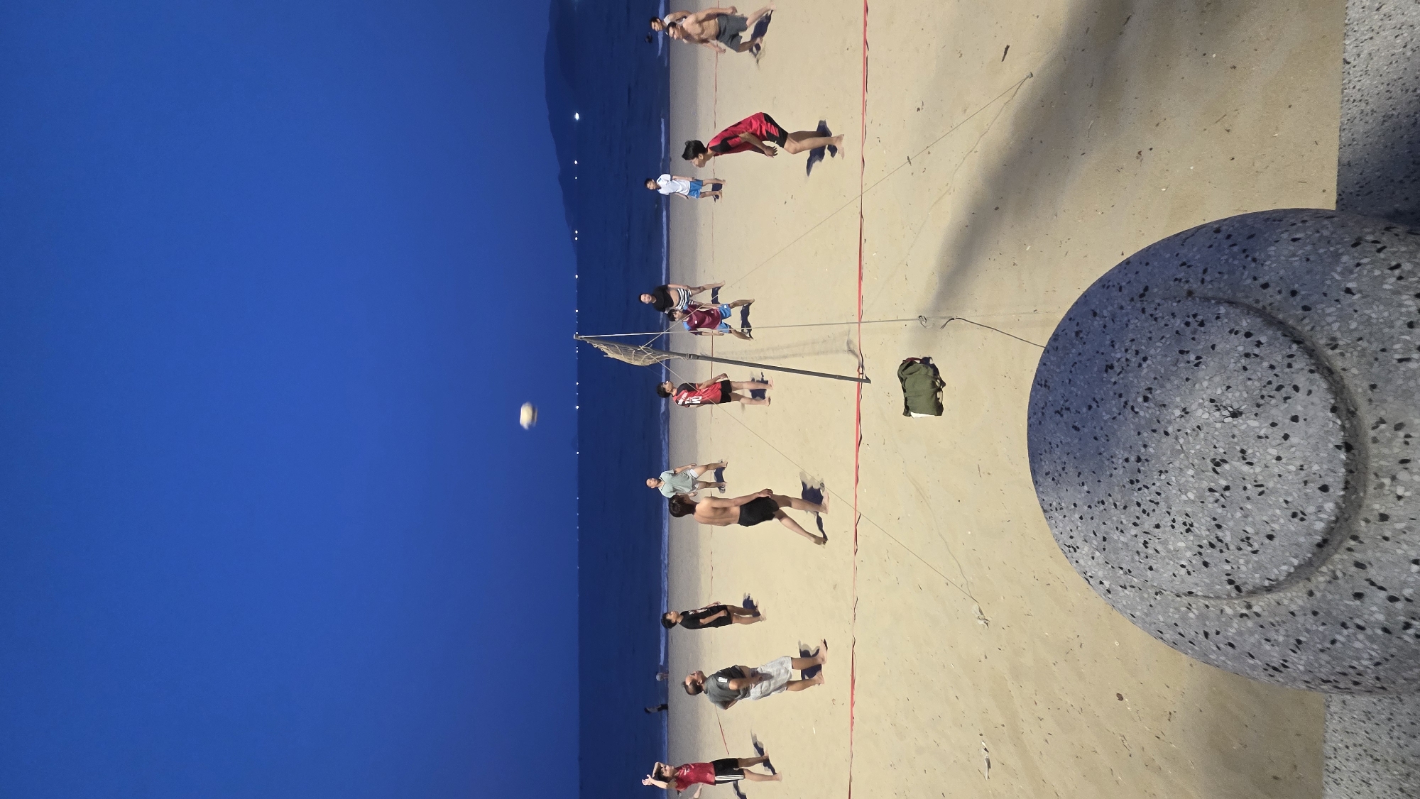 The image shows a beach scene in Nha Trang, Vietnam at dusk. People are playing volleyball near the ocean. The sky is a deep blue, transitioning to a lighter color near the horizon. The beach is sandy and there is a bench made of speckled rock to the right. A green bag sits on the sand near the volleyball net. The volleyball itself is visible in the air above the net. About a dozen people are visible playing volleyball.