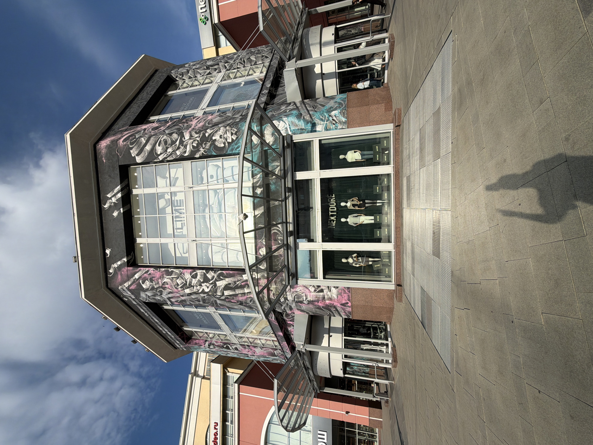 The image shows the exterior of a modern, multi-story building with a distinctive octagonal design. The facade is adorned with a large, stylized mural featuring calligraphic elements in black and pink hues, interspersed with glass windows. On the ground floor, there are several storefronts, including a revolving door entrance and display windows showcasing mannequins dressed in clothing. The foreground is a paved plaza with a geometric pattern of light-colored square tiles. The sky above is blue with scattered white clouds. To the left of the main building, a section of another building with a yellow facade is visible, and to the right, a building with a red facade and arched windows. A shadow of a person holding up their hands is cast on the plaza in the lower right portion of the image.