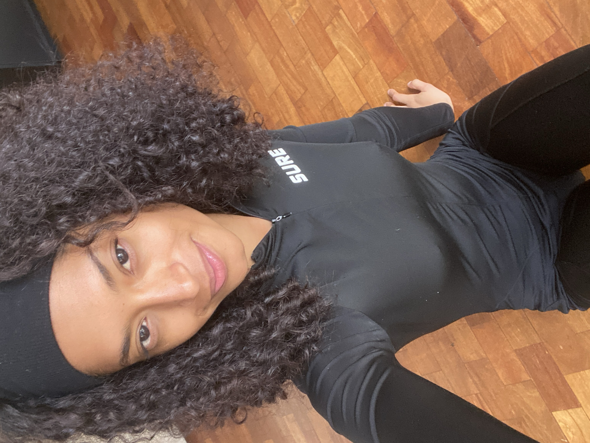 A person with dark, voluminous curly hair and a black headband is pictured lying on a light brown wooden parquet floor. The individual is wearing a form-fitting black long-sleeved top and black leggings. The top features the word "SURE" in white, block letters, oriented vertically on the side/shoulder area.
The person is looking directly at the camera with a calm and slightly smiling expression, their head tilted. The shot is taken from a slightly elevated perspective, looking down. The setting is an indoor space, indicated by the polished wooden floor. No specific time of day or weather conditions are discernible from the image. This scene takes place in São Paulo, Brazil.