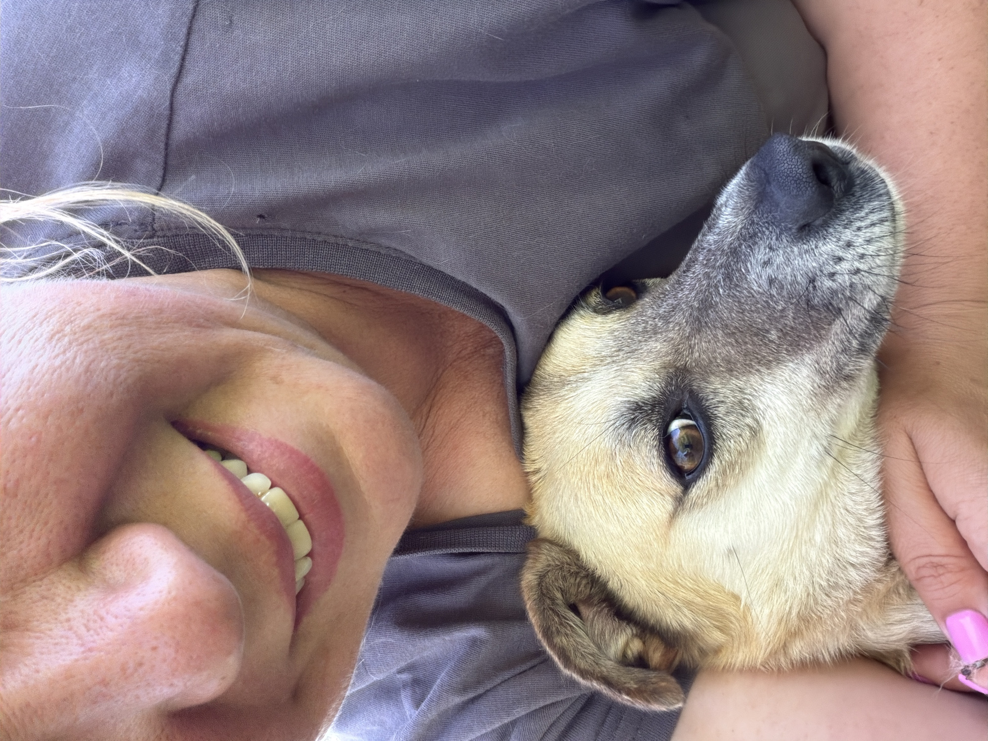 A close-up, slightly angled shot depicts a woman and a dog in an intimate pose. The woman is smiling broadly, showcasing her teeth. Her blond hair is partially visible near her face. The dog, with a light brown and gray coat, rests its head near the woman's chest. The woman's hand is visible, with painted pink fingernails, gently resting on the dog. Both the woman and dog seem to be in close physical contact, suggesting a bond. The woman is wearing a gray top. The background is out of focus, giving no context of a location in Leningradskaya, Russia.