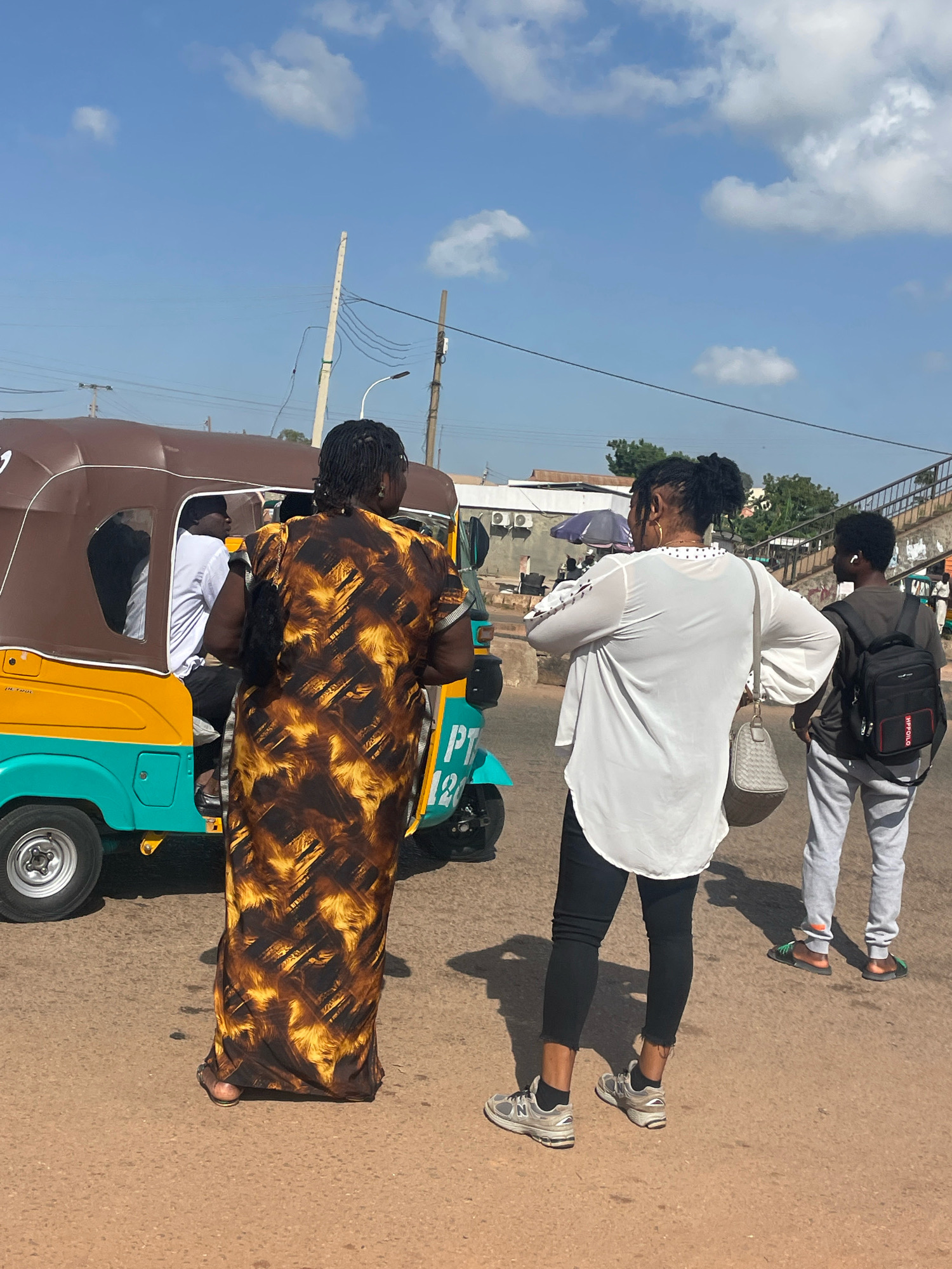 In Gura Top, Nigeria, on a sunlit day, three individuals are visible near a colorful auto-rickshaw. A woman with dark braided hair, wearing a long, patterned brown and yellow dress, stands facing away from the camera, next to the auto-rickshaw. Inside the auto-rickshaw, a man wearing a white shirt is seated. To the right of the woman in the patterned dress, another woman, also with dark hair styled in a bun, is seen from the back. She is wearing a white, loose-fitting top with flared sleeves, black pants, and grey sneakers. A light-colored shoulder bag hangs from her left shoulder. Further to the right, a young person with a black backpack and grey sweatpants is visible from the back, wearing earbuds and sandals. The auto-rickshaw itself is predominantly yellow and turquoise, with brown canvas covering its passenger compartment. The ground is a dusty, reddish-brown surface, indicative of a dirt road. Power lines and poles, along with some buildings and trees, form the background under a bright blue sky with scattered white clouds.