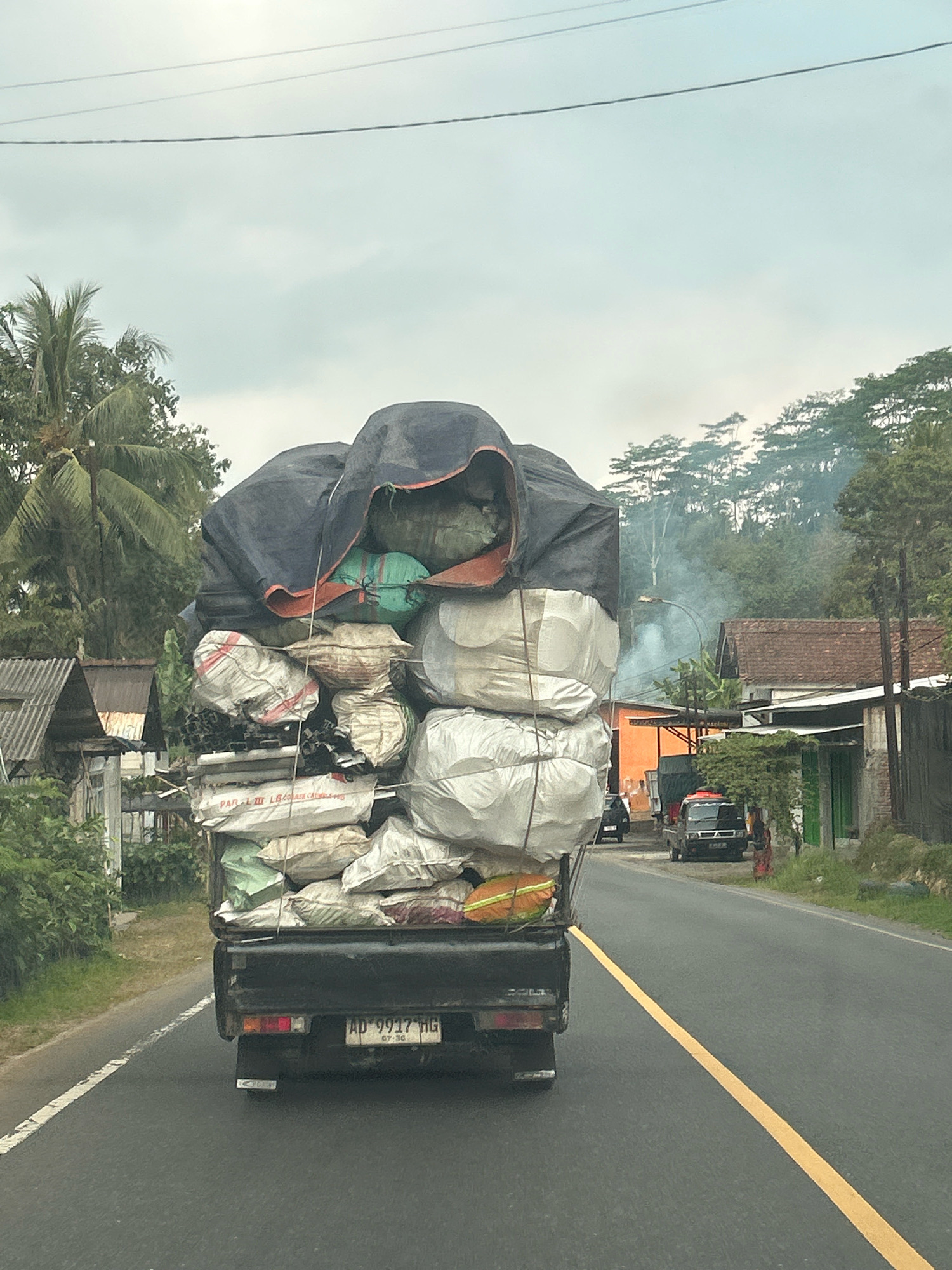 A black pickup truck, license plate AD 9917 HG, is heavily laden with various items covered by a large, dark tarp. The truck is driving on a paved road with a yellow dividing line. To the left of the road, there are trees and a small building with a corrugated roof. To the right, there are more trees, houses with tile roofs, and some vegetation. Smoke is rising in the background, possibly from a distant fire or industrial activity. The sky is overcast. The scene appears to be a rural or semi-rural area in Maja tengah, Indonesia.
