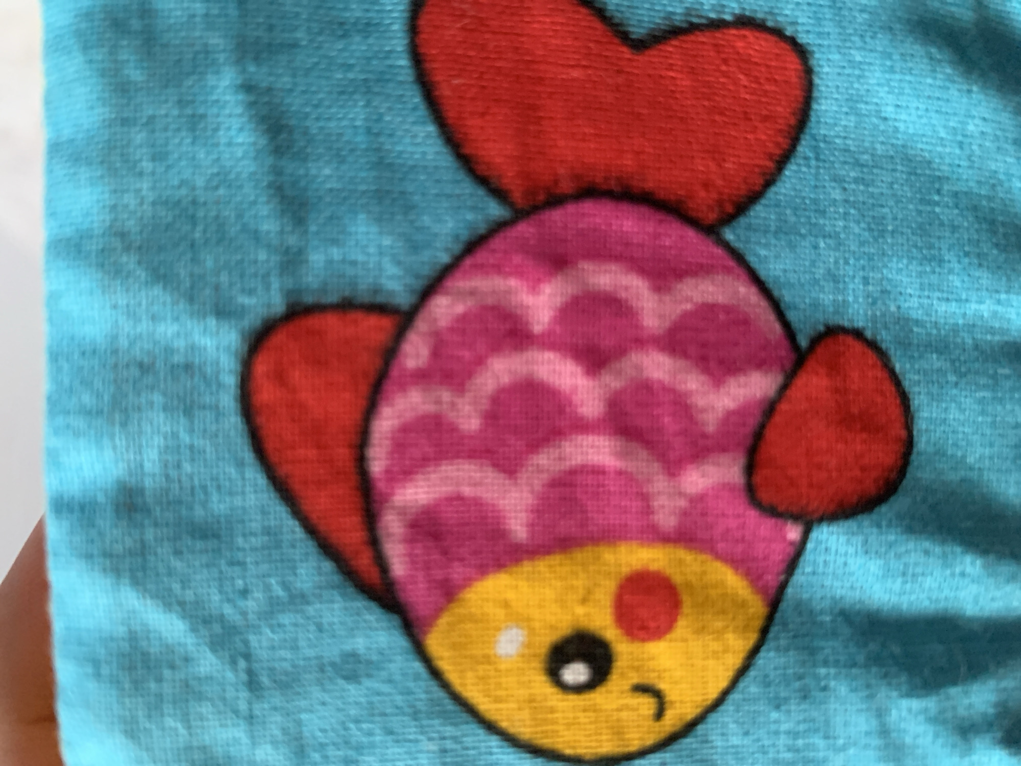 A close-up, top-down shot shows a cartoon fish embroidered on a bright blue fabric. The fish is predominantly pink with a yellow head. Its body is decorated with light pink scale patterns. It has a red tail fin, dorsal fin, and pectoral fin. The fish's face features a black eye, a red circular cheek, and a smiling mouth represented by a black curved line. A small white dot is above the eye, likely a highlight. The fabric has a visible texture of woven threads. The image is framed by a hand holding the fabric from the top left corner. The image does not provide any information about its location in Stavropol, Russia.