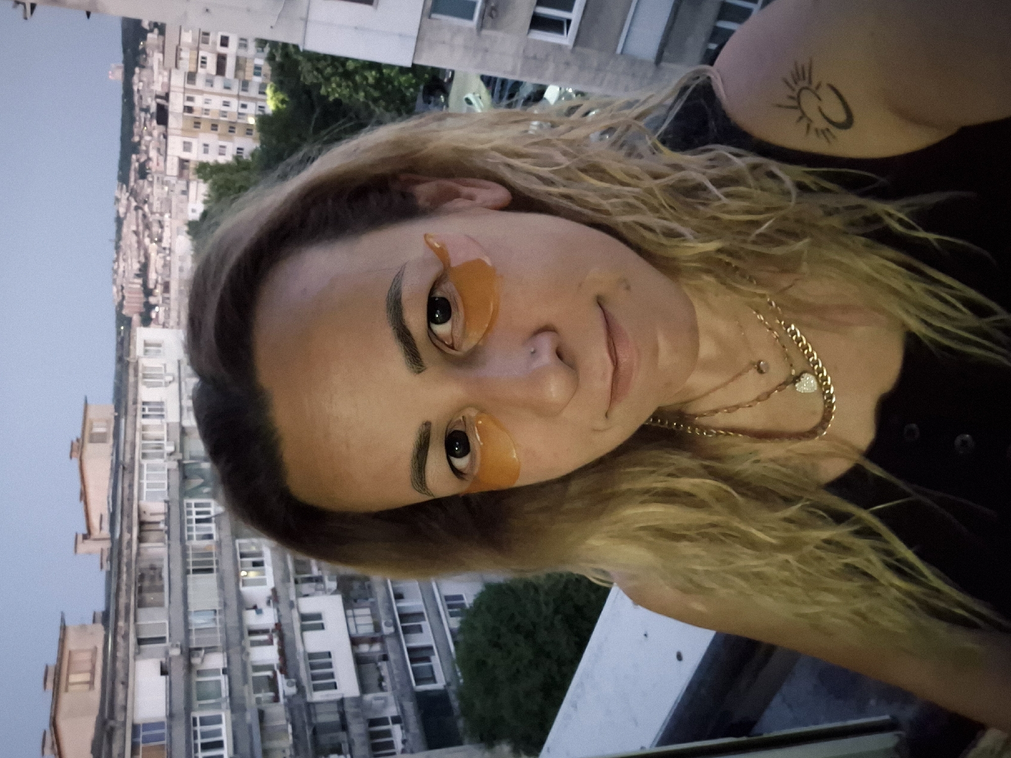 In an outdoor, medium shot, a woman with blonde, wavy hair is standing on a balcony, facing the camera. She is wearing a black tank top and several gold necklaces, one of which has a heart-shaped pendant. She has orange eye patches under her eyes. A black tattoo of a crescent moon and sun symbol is visible on her upper left arm. Behind her, apartment buildings and a cityscape are visible under a twilight sky.