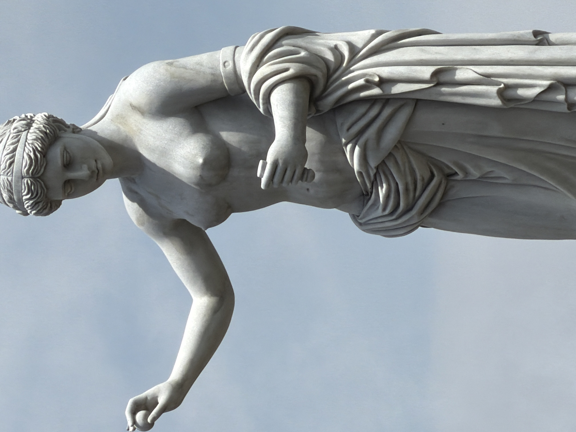 The image depicts a marble statue of a woman against a pale blue sky. The statue is of a classical style with the woman wearing a draped garment. Her hair is styled with a band. She has one arm extended downward, holding a small, round object. Her other hand holds a cylindrical object. The statue is in Đà Nẵng, Vietnam.