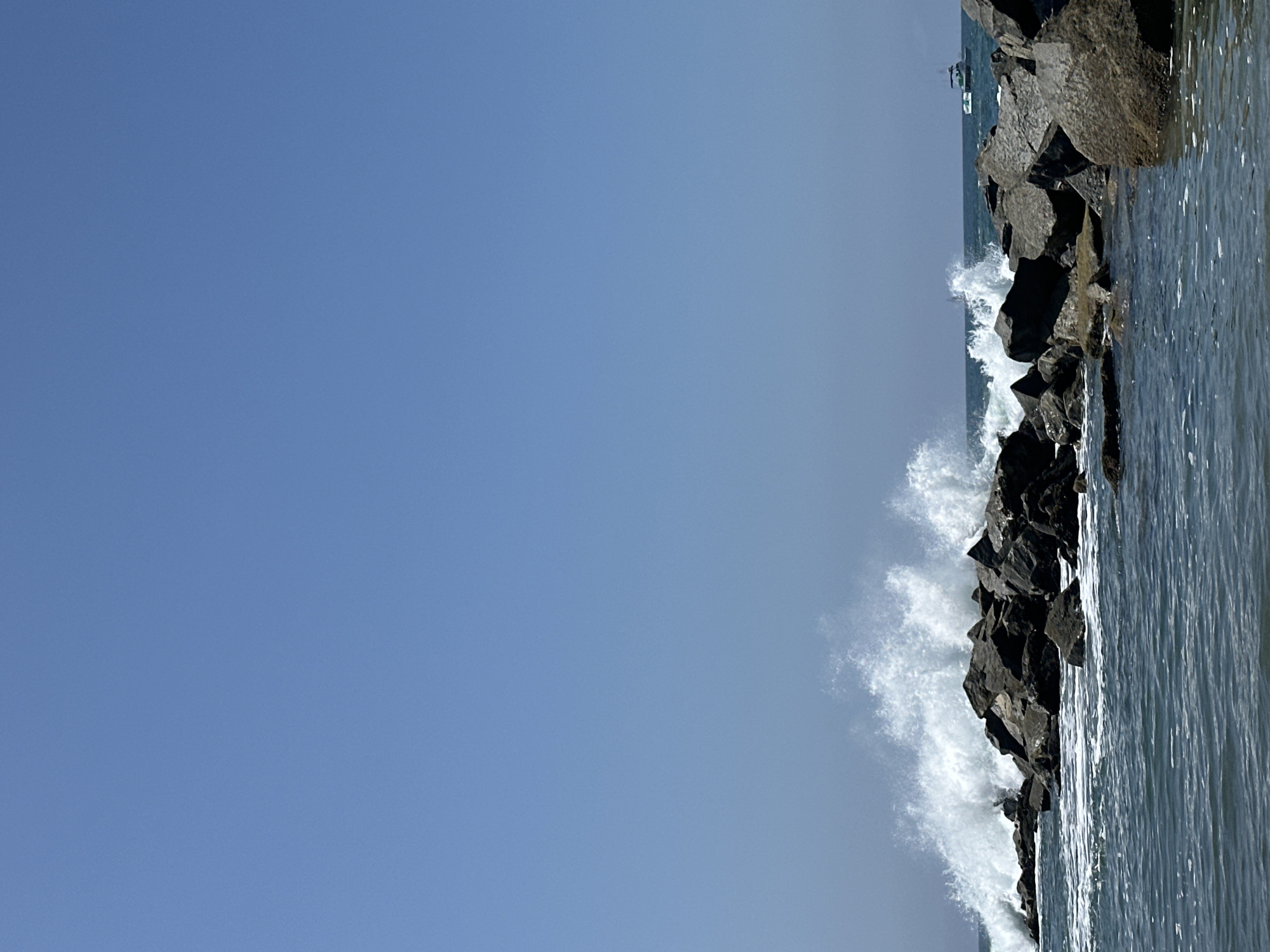 The image shows a rocky breakwater on a clear day with a bright blue sky. Waves are crashing against the rocks, creating white spray. In the distance, two boats are visible on the water. The scene appears to be a coastal area, possibly a beach or harbor. The location in the city of Los Angeles, United States of America, is likely near the ocean, such as the Santa Monica Pier or the Marina del Rey. The water is a deep blue, and the rocks are dark gray and rough-textured. The overall impression is one of a dynamic and natural coastal environment.