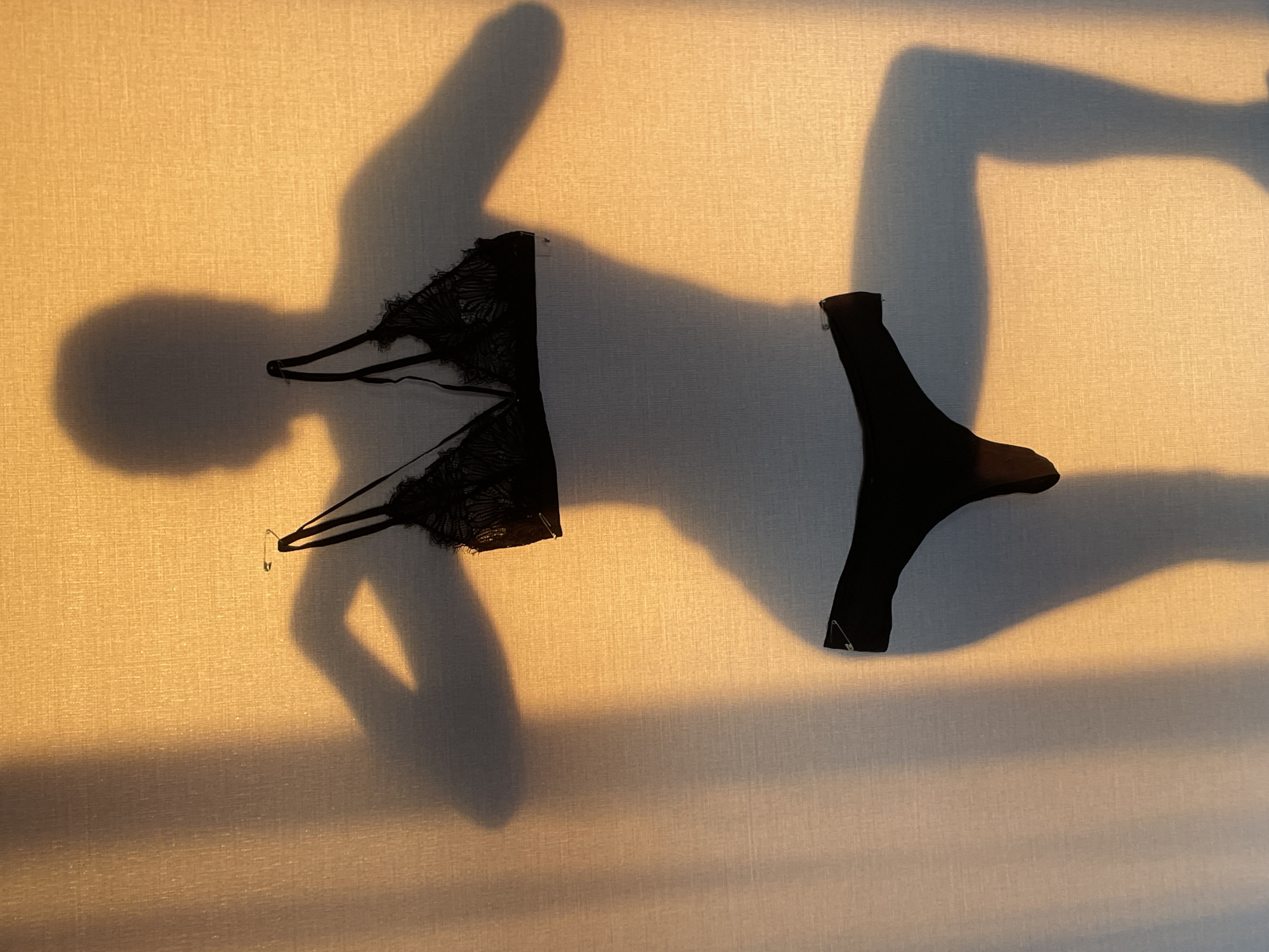 The image shows a bra and underwear pinned to a wall with safety pins. The bra is black lace. The underwear is black. The wall is textured and cream colored. The lighting is uneven, creating strong shadows that give the impression of a person's body surrounding the lingerie. The location, Подшивалово, Russia, cannot be confirmed from the image.