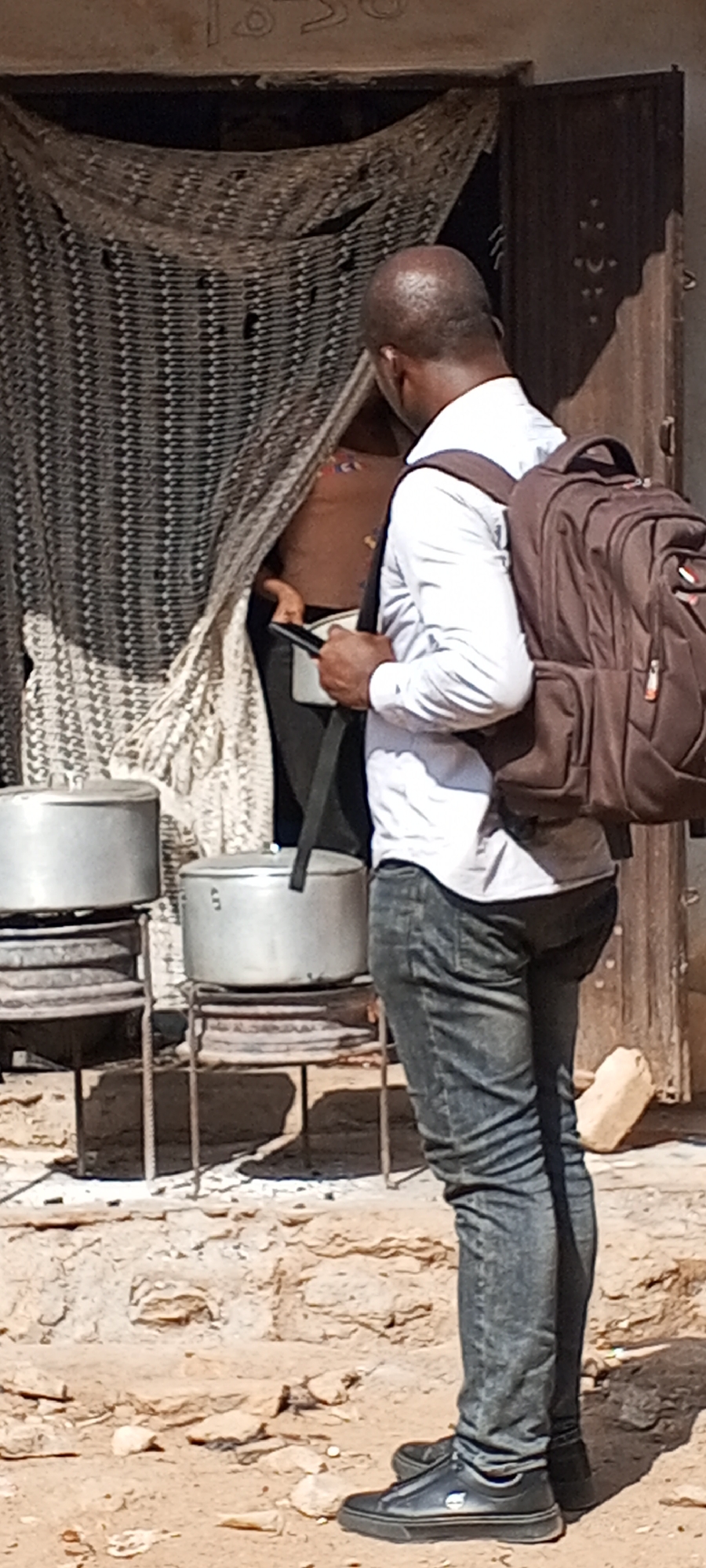 In Sabon Tasha, Nigeria, a man with a shaved head, wearing a light-colored long-sleeved shirt, dark jeans, and black shoes, stands with a brown backpack. He holds a mobile phone in his hand, seemingly interacting with someone partially visible behind a patterned curtain or screen. Two large metal pots rest on stands. The environment appears to be a semi-outdoor space with a building behind the curtain.