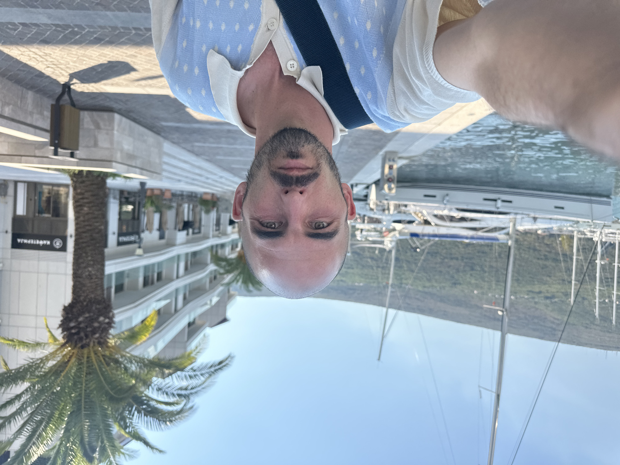 A man with a shaved head, short beard, and light skin is taking a selfie with a harbor in the background. He is wearing a light-colored sweater with blue diamond patterns and a dark blue strap across his chest. The harbor features several sailboats and a modern building with balconies and palm trees in front. The scene appears to be in Herceg Novi, Montenegro, with a promenade and paved walkway visible.