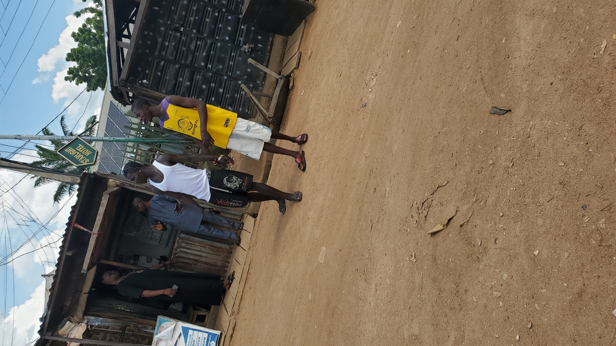 A medium shot captures an outdoor scene in Jalingo, Nigeria, under a cloudy sky. In the foreground, a dirt path is visible. To the left, a man in a black robe stands near a stall with a corrugated metal roof and walls, which appears to be a shop. Behind him, another man in a grey tunic and trousers holds a smartphone and gestures with his hand. In the center and right of the frame, two young men stand close together. One, in a white tank top and black shorts with skull graphics, faces left. The other, wearing a yellow tank top with a graphic and white shorts, faces slightly right. Behind them, the "STARLIGHT HOTEL" sign is visible on a green placard, affixed to a pole. Further back, structures with dark, corrugated metal siding are present, along with some wooden elements. Palm trees are visible in the background, suggesting a tropical or sub-tropical environment.