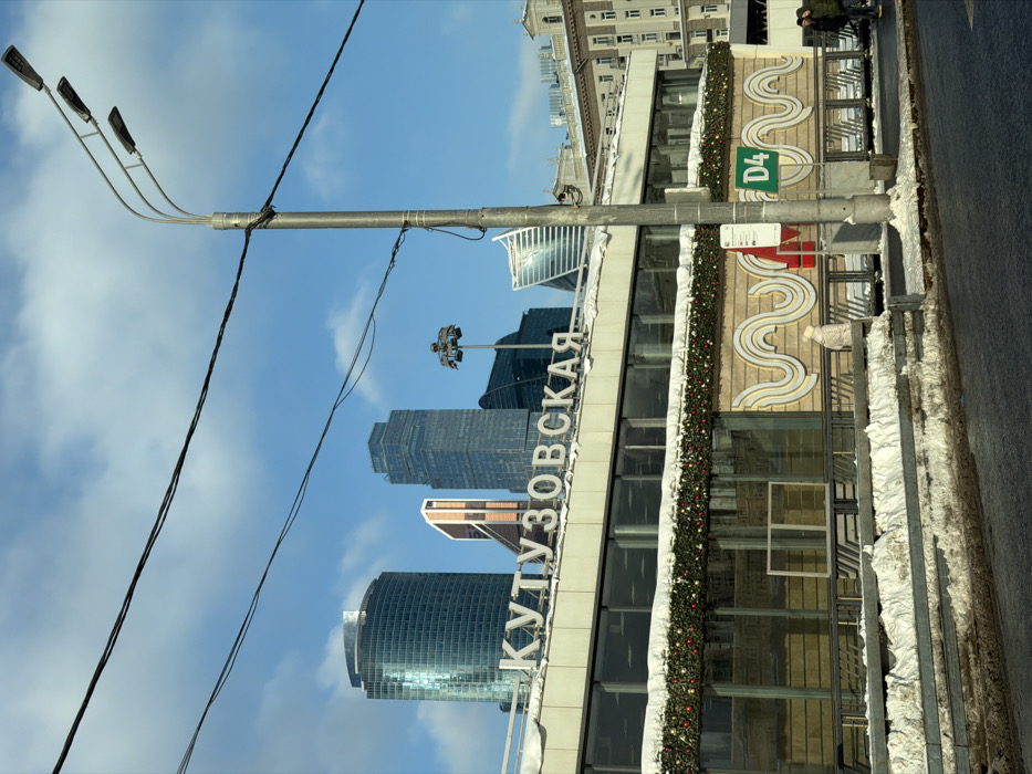 The image depicts a winter urban street scene at the Kutuzovskaya area in Moscow, Russia, under a clear blue sky with scattered white clouds. In the foreground, snow and slush cover the ground along a paved street. A grey metallic utility pole with two horizontally extended streetlights stands on the right, with dark utility wires crisscrossing the upper portion of the frame. A green square sign displaying "D4" in white is affixed to the pole, alongside a smaller red and white sign.
The midground features a two-story building, likely a transport station entrance, with "КУТУЗОВСКАЯ" (KUTUZOVSKAYA) prominently displayed in large white Cyrillic letters along its roofline. The building's upper level features large rectangular windows, while the lower façade is adorned with a decorative wavy pattern in beige and white tones. A festive garland, composed of green foliage and red and gold ornaments, is draped across the building below the windows, partially covered in snow. A thin layer of snow also rests on the building's roof. A single individual wearing light-colored winter clothing walks on the pavement in front of this building, near the wavy façade. To the far right, two additional individuals in dark winter attire are visible walking on the pavement.
In the background, a cluster of modern skyscrapers from the Moskva-City business district is visible, including a cylindrical glass tower, a tall rectangular dark glass building, and a distinctive copper-paneled structure. Further to the right, a classic multi-story building with a light-colored façade and numerous windows provides architectural contrast.