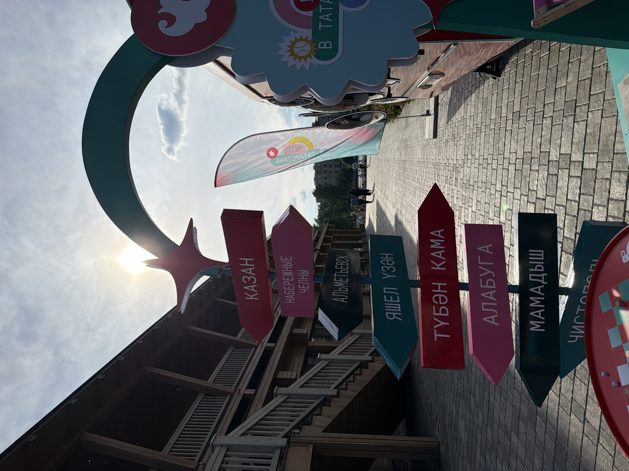 The image depicts an outdoor scene with a directional signpost in the foreground, featuring multiple colorful arrows pointing in different directions with city names written in Cyrillic script. The names visible include "КАЗАН", "НАБЕРЕЖНЫЕ ЧЕЛНЫ", "АЛЬМЕТЬЕВСК", "ЯШЕЛ ҮЗӘН", "ТУБӘН КАМА", "АЛАБУГА", "МАМАДЫШ", and "ЧИСТОПОЛ". Behind the signpost, a wooden building with a visible staircase is present. To the right of the signpost, a red vehicle, possibly a food truck or trailer, is parked, adorned with decorative elements. A large, teal-colored archway is situated above the signpost, and a colorful flag is displayed near the vehicle. The ground is paved with stone tiles, and a few people are visible walking in the background. The sun is shining brightly, casting shadows and creating a lens flare. The setting appears to be a public space, possibly a tourist area or a festival, within the city of Kazan, Russia, given the prominent signage.