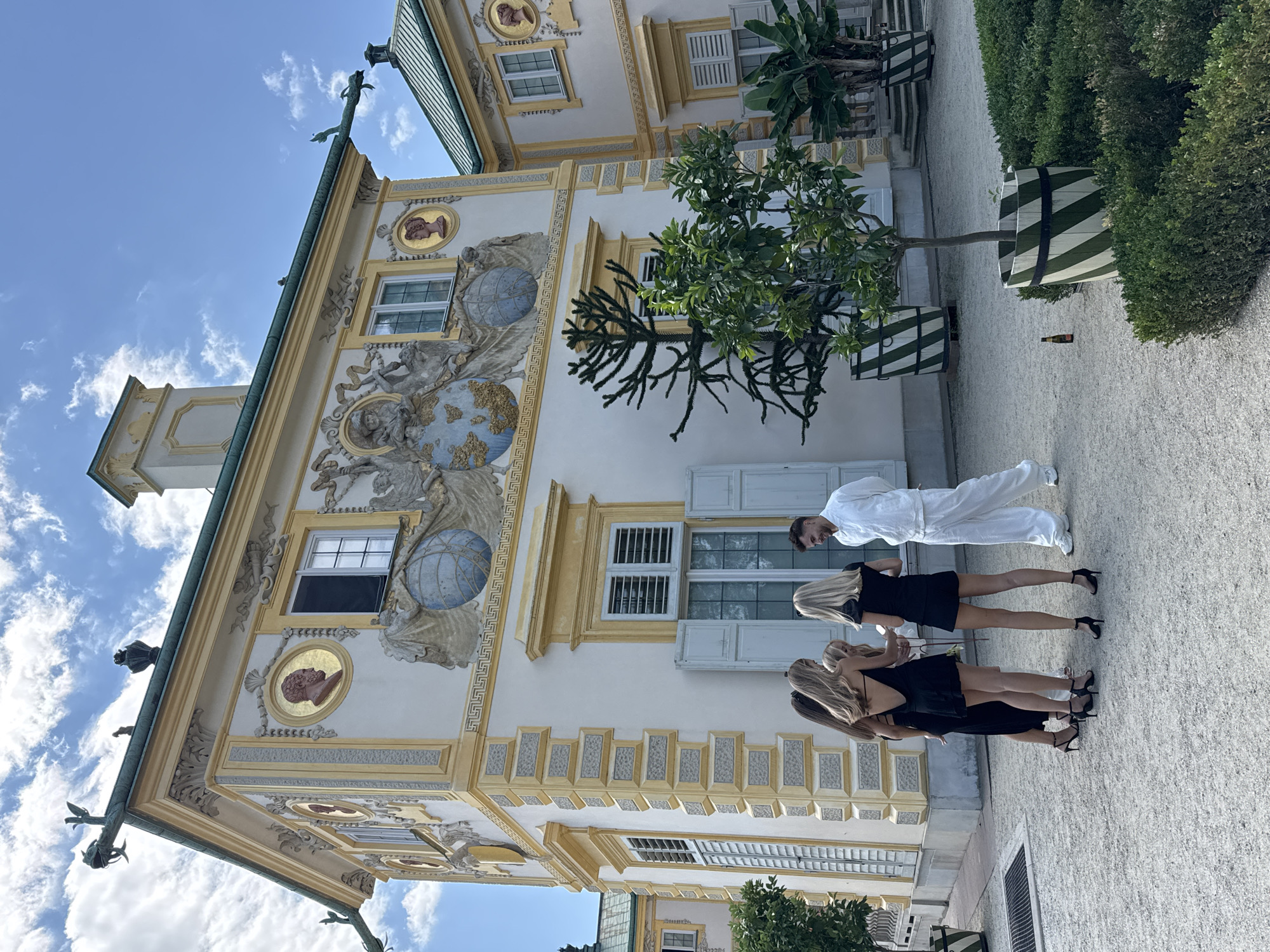 The image depicts a bright, sunny day with a clear blue sky and scattered white clouds. In the foreground, four individuals are gathered in a courtyard paved with light-colored gravel. Three women, dressed in short black dresses and high heels, are facing away from the camera, engaged in conversation. A man, dressed in a white tracksuit, stands facing the women and smiling.
Behind the group, an ornate, cream-colored building with yellow architectural details stands prominently. The building features a green roof and is adorned with decorative elements, including circular portraits in gold frames, elaborate sculptures, and architectural molding. Windows with light blue shutters are visible on the facade.
To the right of the building, large planters with green and white striped patterns contain trees and foliage. The courtyard also has manicured green hedges in the foreground. A small bottle of beer lies on the gravel near one of the planters.
The architectural style and distinct yellow and cream color scheme, along with the overall atmosphere, are characteristic of Wilanów Palace in Warsaw, Poland, a former royal residence known for its Baroque architecture and gardens.