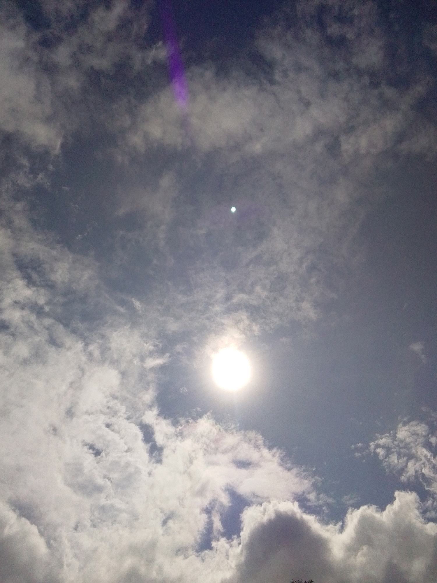 The image captures a dynamic sky over Serti, Nigeria, characterized by a mix of dense and scattered clouds against a deep blue backdrop. The sun is a prominent feature, shining brightly in the lower half of the frame, with a distinct halo effect indicating its intensity. A smaller, less luminous celestial body, possibly the moon or another distant object, is visible higher in the sky. A striking purple light streak cuts diagonally across the upper left portion of the sky, adding an unusual atmospheric element to the scene. The clouds vary in texture, with some appearing wispy and thin, while others are more voluminous and textured, casting subtle shadows. The overall impression is one of a weather-filled sky, perhaps preceding or following a change in conditions.
