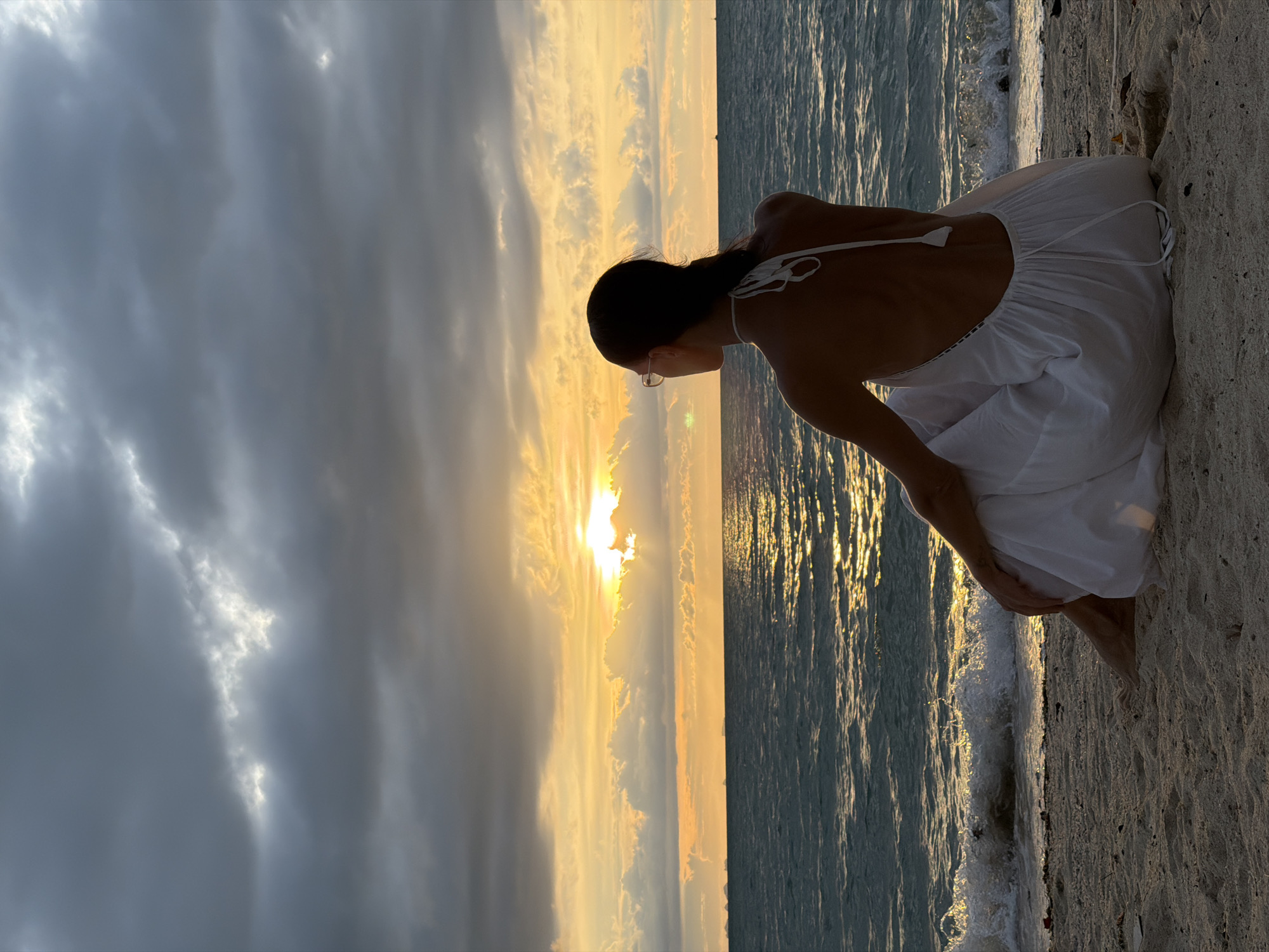 The image depicts a person kneeling on a sandy beach, facing the ocean during sunset. The person is wearing a white dress and is viewed from behind, with their back to the camera. Their hair is pulled back, and they appear to be looking out at the water and sky. The sunset creates a dramatic sky with warm, golden light illuminating the clouds and reflecting on the water. Some clouds are dark and overcast, while others are bright and tinged with yellow and orange. The ocean waves are gently lapping at the shore. The overall mood of the image is serene and contemplative, with the natural beauty of the sunset setting a peaceful tone. There is no visible text in the image.