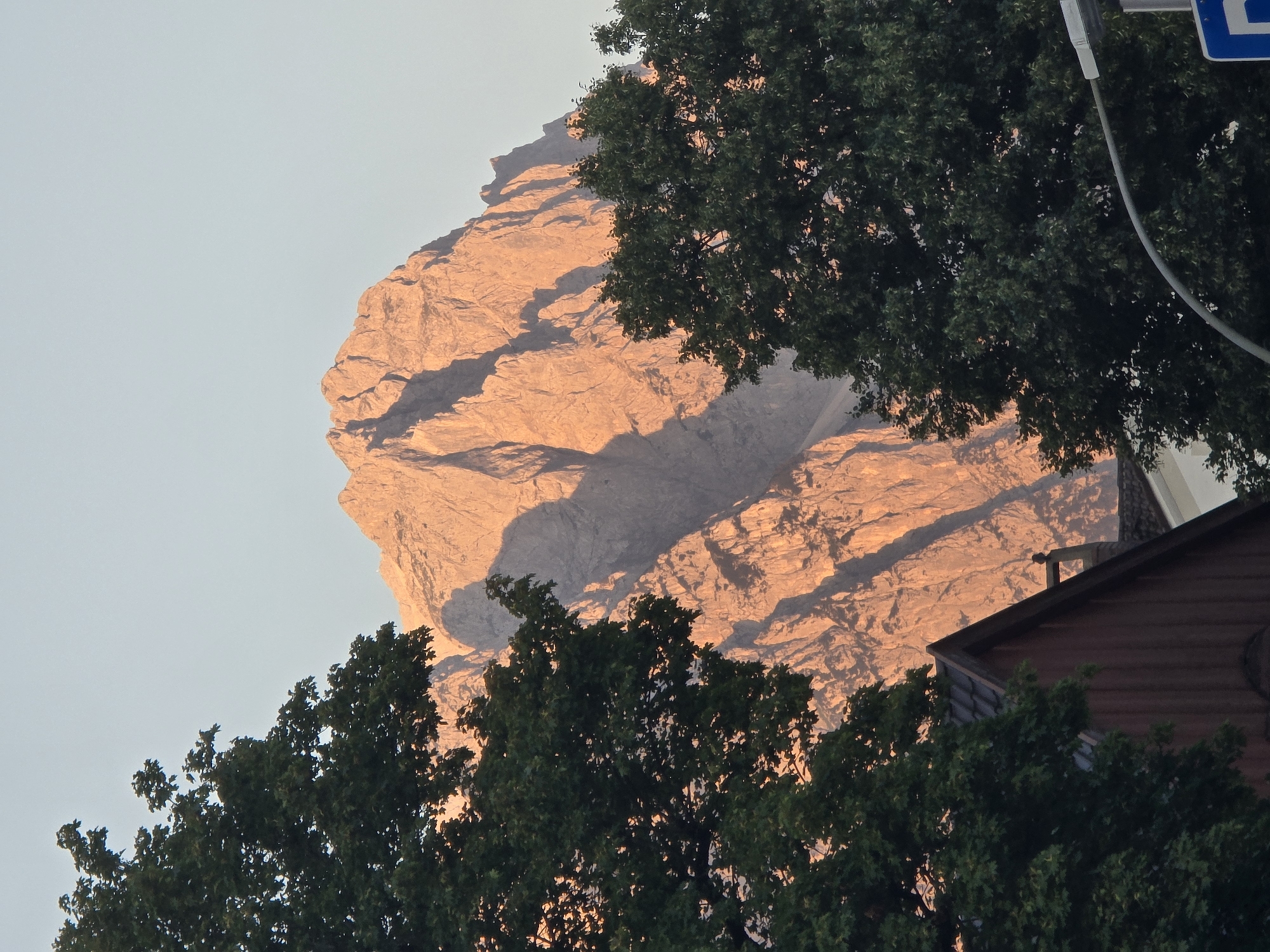 This photograph captures a mountainous landscape with a peak illuminated by the warm light of either sunrise or sunset. The mountain's rugged texture is visible, with sharp ridges and deep shadows. In the foreground, two large, dark green trees frame the view of the mountain. Below the trees, the roofline of a building, possibly a house with a dark wooden facade, is visible. The sky above is a clear, pale blue, indicating fair weather. The scene appears to be located in or near Garmisch-Partenkirchen, Germany, a region known for its scenic Alpine vistas.