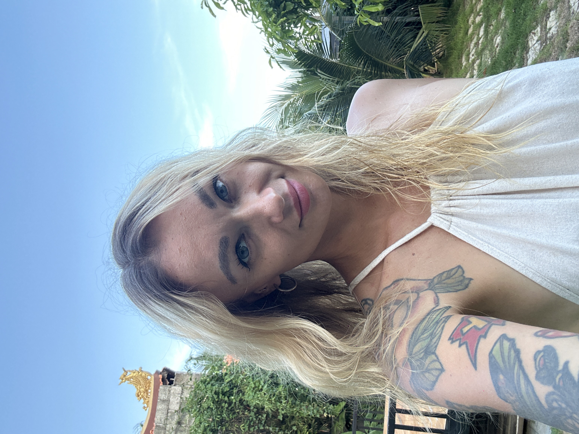 A vibrant outdoor selfie features a young woman with blonde, wavy hair and striking blue eyes, captured in Mũi Né, Vietnam. She looks directly at the camera with a calm, gentle smile, her face framed by her light hair. She wears a light-colored, possibly off-white or beige, halter-neck top. A prominent and colorful tattoo sleeve covers her left arm, showcasing various designs including green leaves, red shapes, and a yellow lightning bolt. She also has a subtle nose piercing and an earring.
The background depicts a bright, sunny tropical setting. A large expanse of clear blue sky fills a significant portion of the upper frame, indicating good weather. Lush green foliage, including the fronds of palm trees, is visible on the right side and behind her, suggesting a rich natural environment. In the lower-left corner, a detail of a traditional architectural structure with an ornate golden element, possibly part of a temple roof or local building, adds a cultural accent to the scene. The overall impression is one of relaxation in a warm, exotic location. No text is visible in the image.