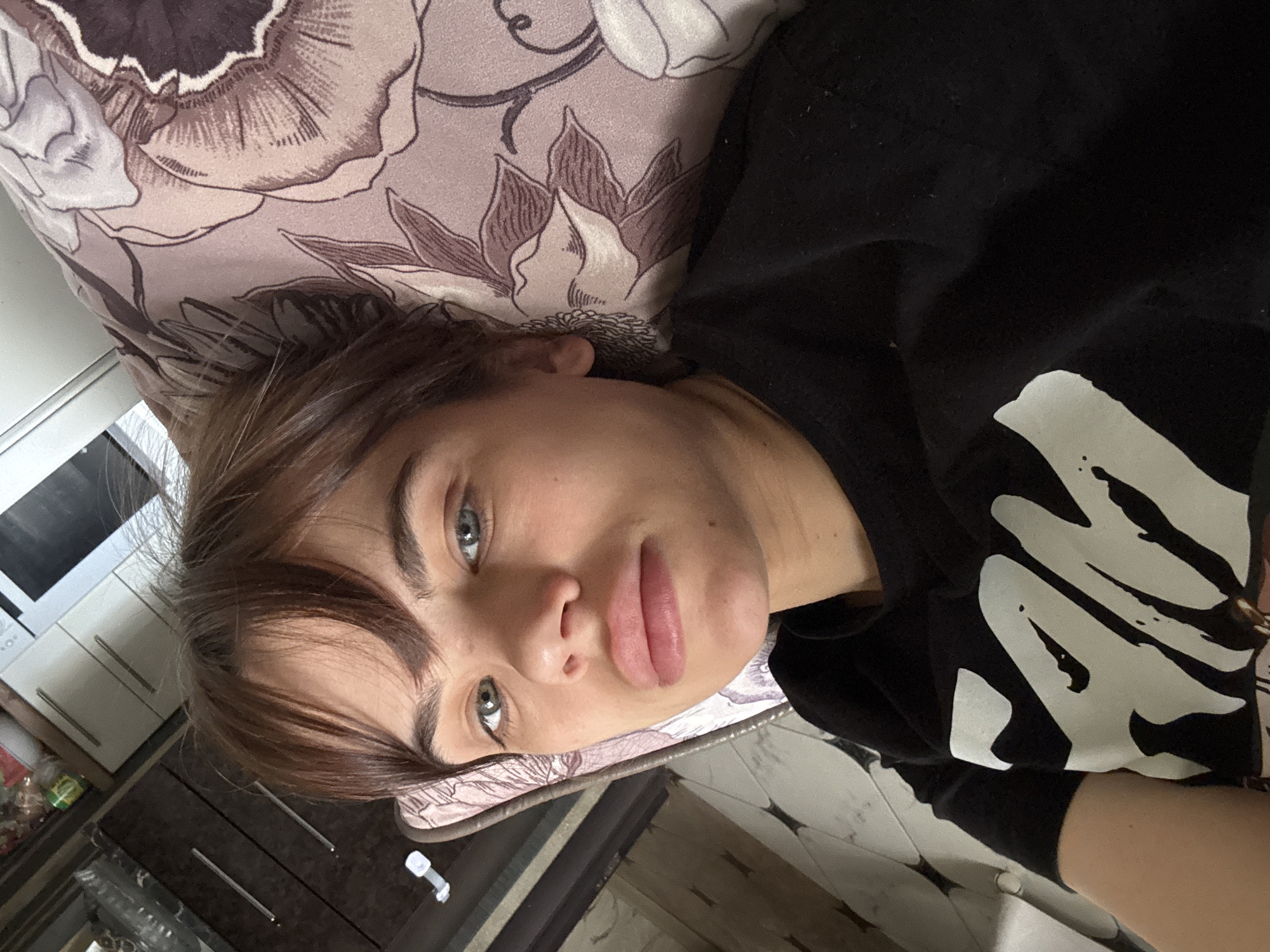 A young woman is depicted in a close-up, tilted shot, lying down indoors. She has striking light blue/gray eyes, brown hair with a fringe, and full lips, with a neutral and calm expression. She is wearing a black t-shirt featuring a large, abstract white graphic design that covers a significant portion of the fabric.
Behind her head, a pillow or blanket with a muted floral pattern in shades of purple, brown, and cream is visible. In the background, out of focus, are household furnishings including white cabinets, a microwave, and darker cabinets with silver handles, suggesting a domestic interior. The setting appears to be a room, possibly a bedroom or living area. The image is a selfie or portrait taken from above, capturing her relaxed pose while lying down. The location context is Rostov-on-Don, Russia. No discernible text is visible, only the graphic pattern on the shirt. Time of day and weather are not evident.