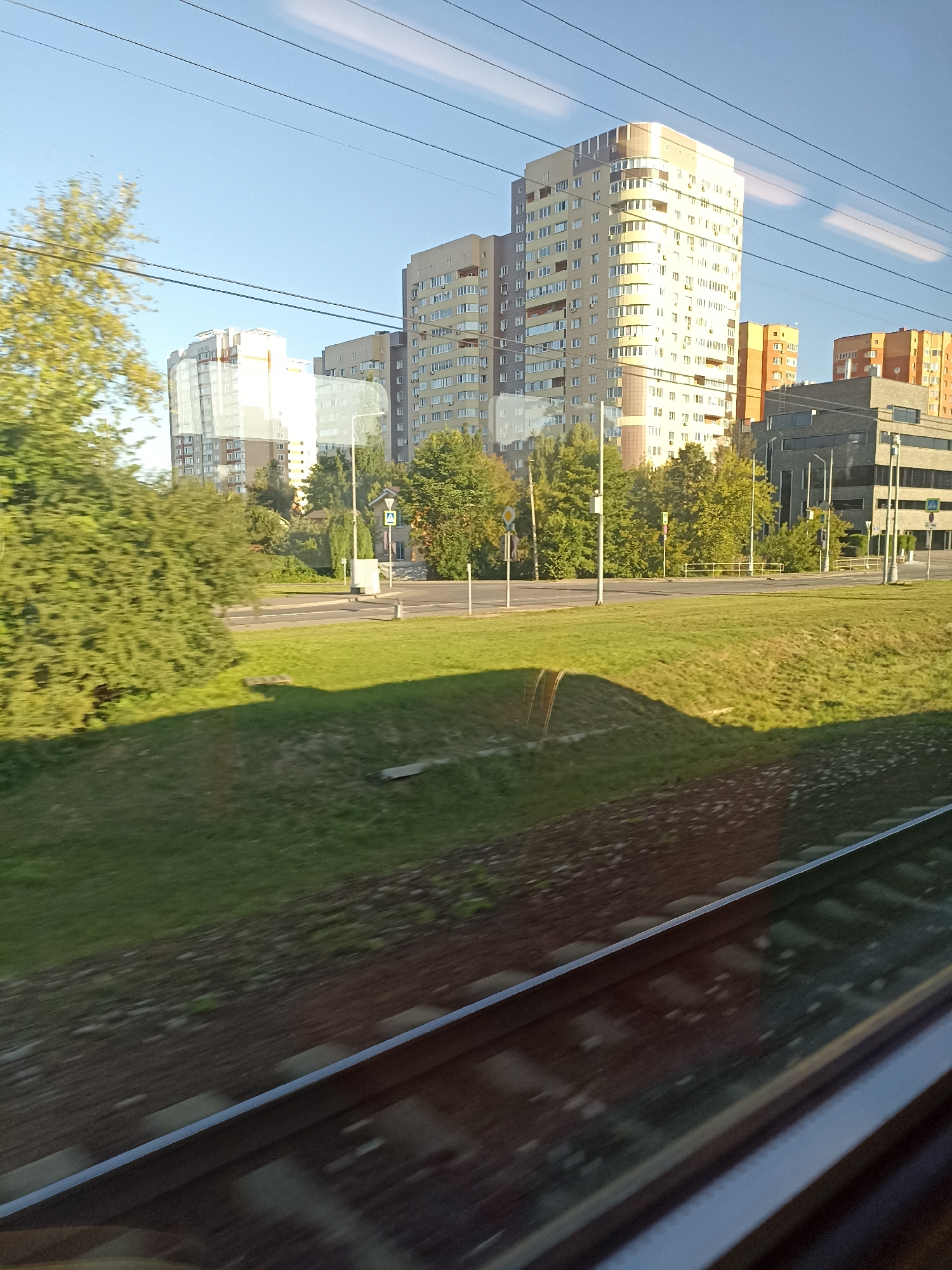 This image shows a view from a train window, looking out at a city scene in Shcherbinka, Russia. The foreground features train tracks running diagonally from the bottom left to the bottom right. Beyond the tracks, there is a grassy embankment. In the middle ground, a road with a crosswalk and traffic lights is visible, with trees and a few small buildings lining it. The background is dominated by several multi-story residential buildings, with one particularly tall, light-colored building in the center. The sky is clear and blue, with some light clouds. The overall impression is of a suburban or residential area of a Russian city.