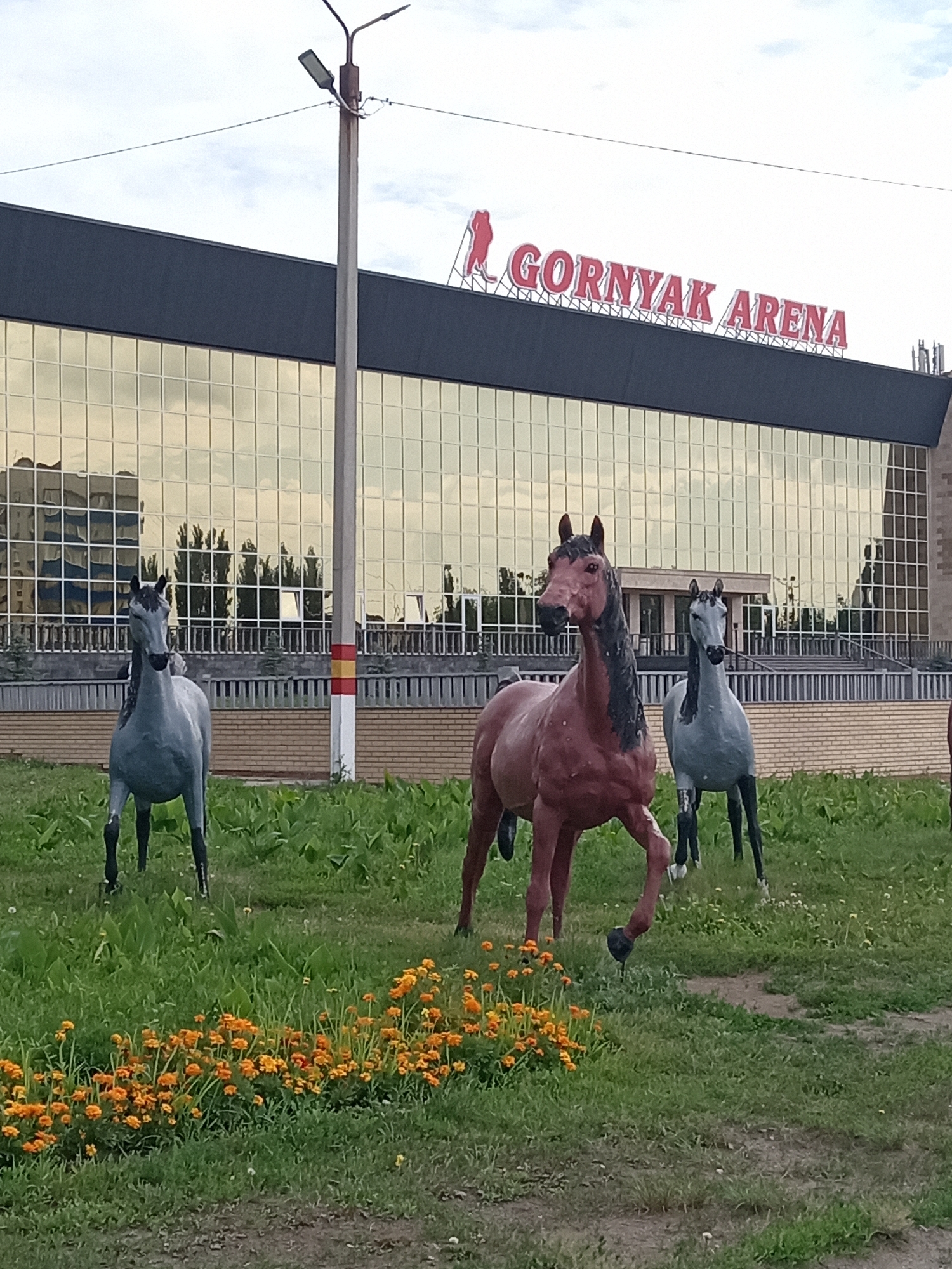 The image shows a bright, overcast day in Rudny, Kazakhstan, in front of the GORNYAK ARENA. Three life-sized statues of horses are positioned in a grassy area in the foreground, with bright orange flowers blooming to the left. The GORNYAK ARENA is a large building with a dark facade and extensive gold-tinted windows, reflecting the sky and surrounding trees. A street light stands prominently to the left of the building. The horses are depicted in various poses, with one in the center appearing to trot. The overall scene is static, with no people visible.