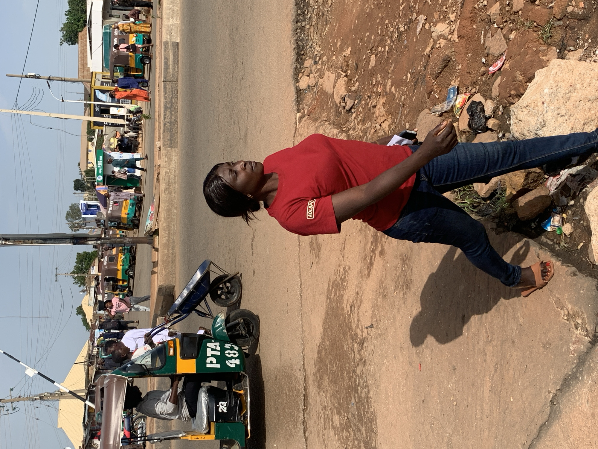 In Gura Top, Nigeria, a woman in a red t-shirt and blue jeans walks on a dirt path beside a street. The woman is carrying a white object in her right hand. In the background, several auto-rickshaws are parked on the side of the road, and a few people are visible near the vehicles and in front of small shops. The environment is dusty with sparse vegetation and scattered rocks. The scene appears to be a busy street or market area.