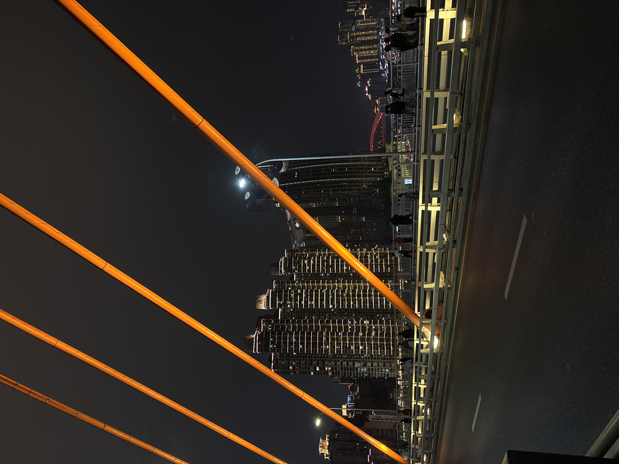 A nighttime photograph captures a striking urban landscape from a bridge. The image is taken in portrait orientation, with prominent orange suspension cables extending diagonally from the lower left corner towards the upper right. To the right of these cables, a dark asphalt road features a dashed white line. Further right, a white metal railing, brightly illuminated by integrated lights, borders the bridge.
Beyond the railing, a sprawling cityscape unfolds with numerous high-rise buildings densely packed and sparkling with countless lit windows. In the mid-ground, several silhouetted figures of people are visible on the bridge behind the railing, appearing to stand or walk, enjoying the night view. A distinctive red arched structure can be seen illuminated in the distance among the city lights. The dark night sky is visible above the buildings, with a faint, glowing orb, possibly the moon, barely discernible.
The scene depicts a bustling, modern metropolis at night, likely in Yuzhong District, China, characterized by its impressive architecture and vibrant lighting. The time of day is clearly night, and the weather appears clear. No visible text is present in the image.