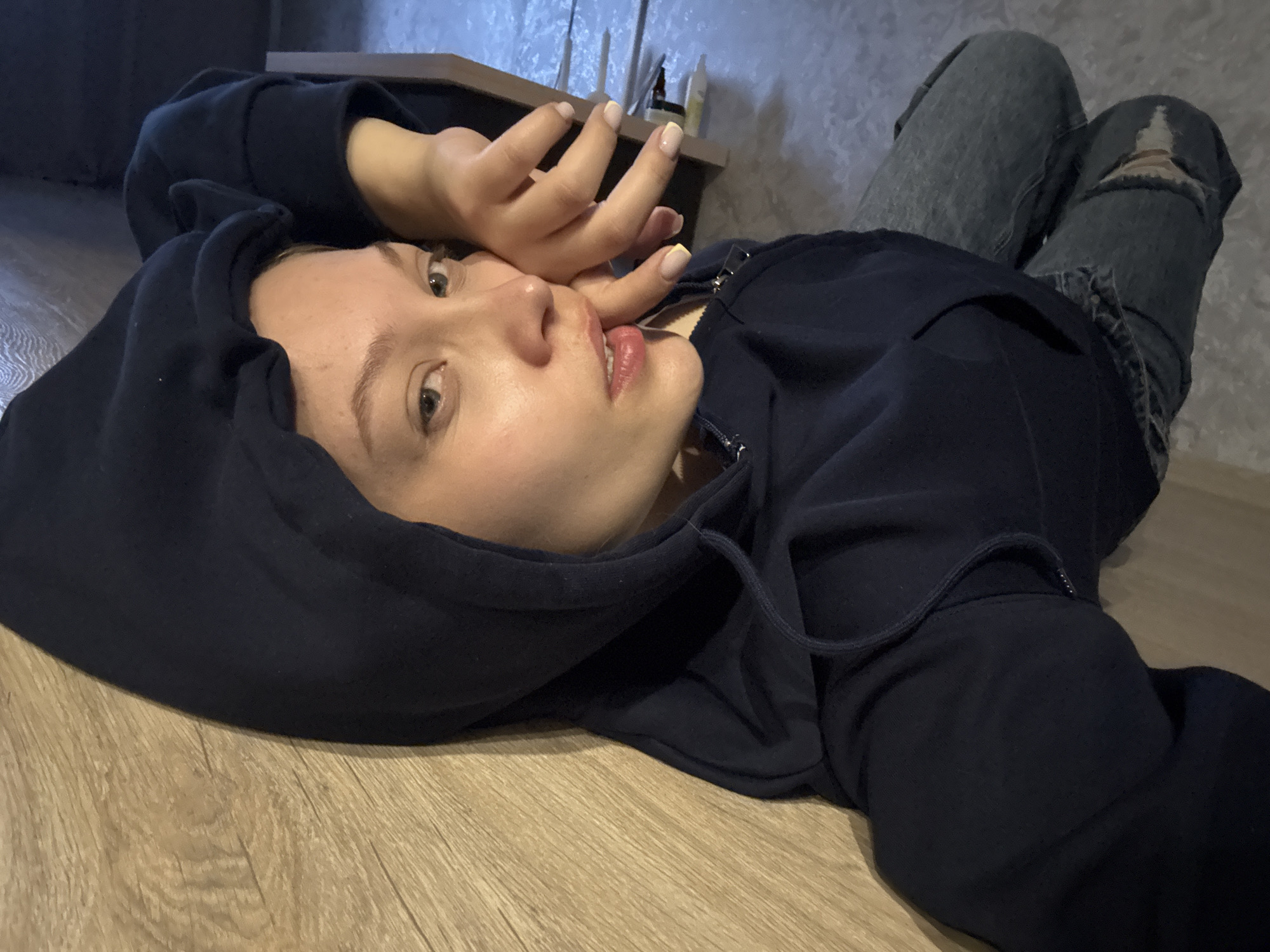A young woman wearing a dark blue hooded sweatshirt and ripped blue jeans is lying on a wooden floor. She is looking upwards, with her right hand near her face. Behind her is a shelf with various objects. The photograph appears to have been taken indoors. The location is Chaikovsky, Russia.