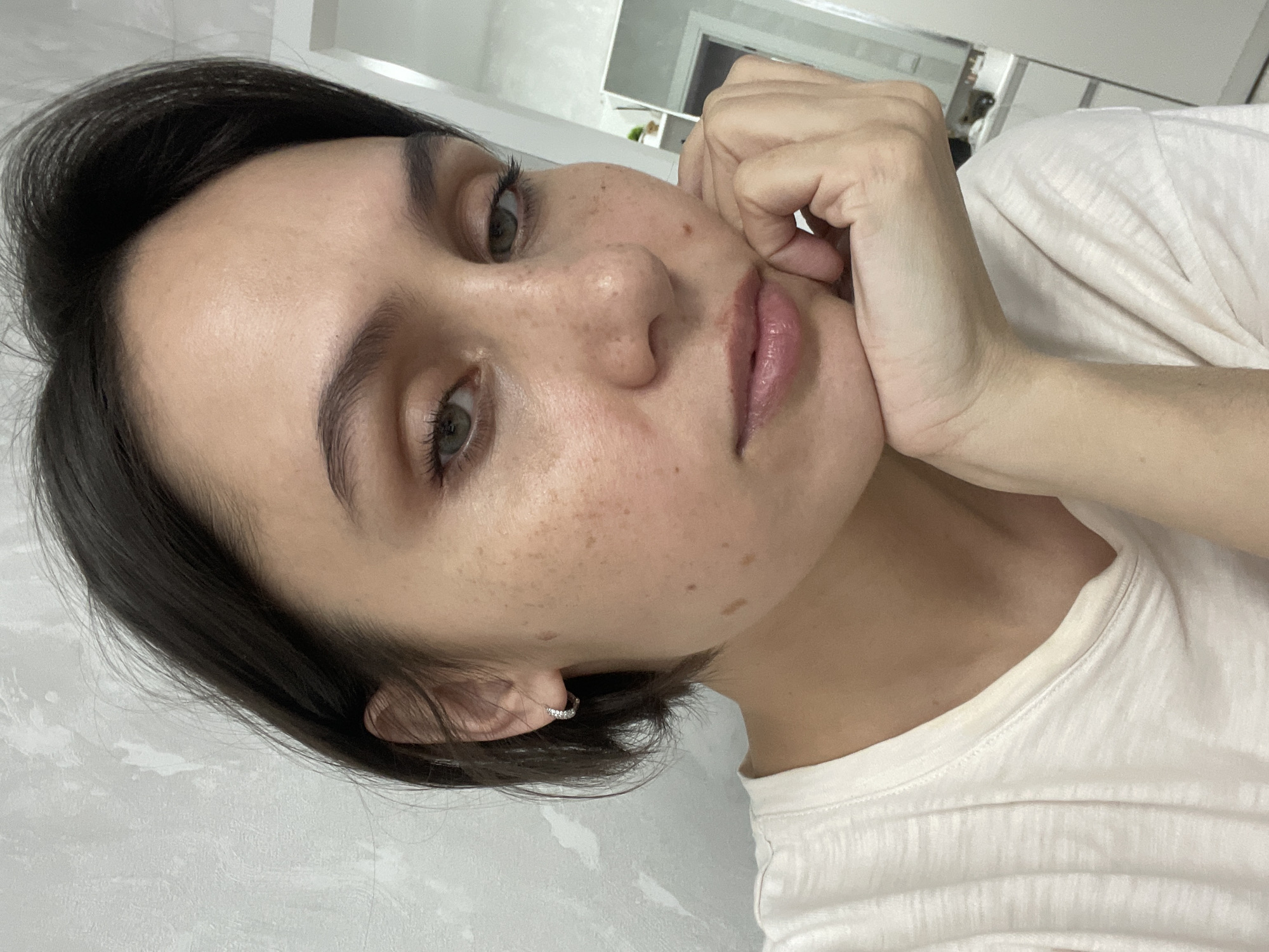 A close-up, slightly tilted portrait shows a young woman indoors in what appears to be Izhevsk, Russia. She has short, dark brown hair that frames her face, fair skin with numerous freckles across her nose and cheeks, and striking green eyes. Her eyebrows are dark and well-defined, and she has visible eyelashes. Her lips are a light, natural pink hue with a glossy finish.
She is wearing a simple white t-shirt. Her right hand is propped under her chin, with her fingers curled around her jawline, suggesting a thoughtful or relaxed pose. A small, sparkling hoop earring is visible in her left ear. She gazes directly at the camera with a neutral to slightly contemplative expression.
The background is minimalist, featuring a light gray or white textured wall. In the upper right portion of the image, a mirror reflects a brightly lit room, indicating an indoor setting, possibly a bathroom or dressing area, characterized by clean lines and light colors. The lighting is soft and even, suggesting either diffused natural light or artificial indoor lighting, making it difficult to determine the exact time of day, though it appears to be bright. No text is visible in the image.
