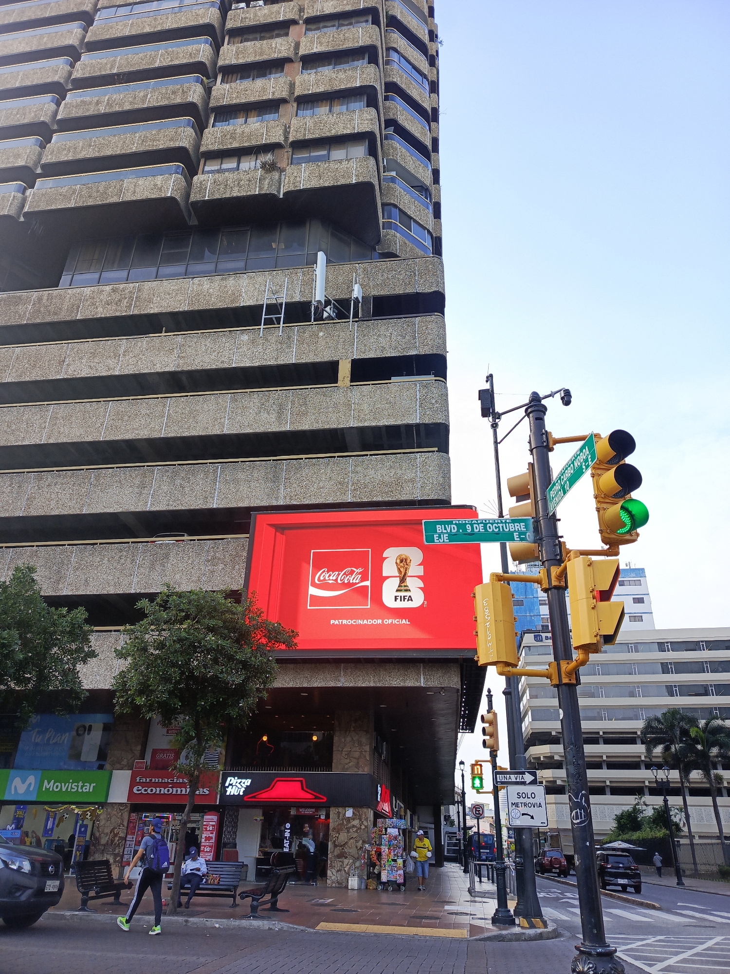 The image captures a street scene in Guayaquil, Ecuador, dominated by a tall, Brutalist-style building with cantilevered balconies. At the base of the building, a large red digital billboard displays a Coca-Cola advertisement with a FIFA World Cup graphic. To the right of the billboard, a traffic light pole stands with multiple traffic signals, including a green light illuminated, and street signs indicating "ROCAFUERTE BLVD. 9 DE OCTUBRE EJE" and "PEDRO CARBO NOBOA AVENIDA 10 S-E".
The sidewalk is bustling with activity. A person wearing a blue cap, backpack, and athletic wear walks from left to right. Across the street, several cars are visible, including a black SUV and a red sedan. Palm trees line the side of the street, and other buildings, some with modern architecture, are in the background. Storefronts like "Movistar," "Farmacias Economicas," and "Pizza Hut" are identifiable along the street level. The sky is clear and blue.